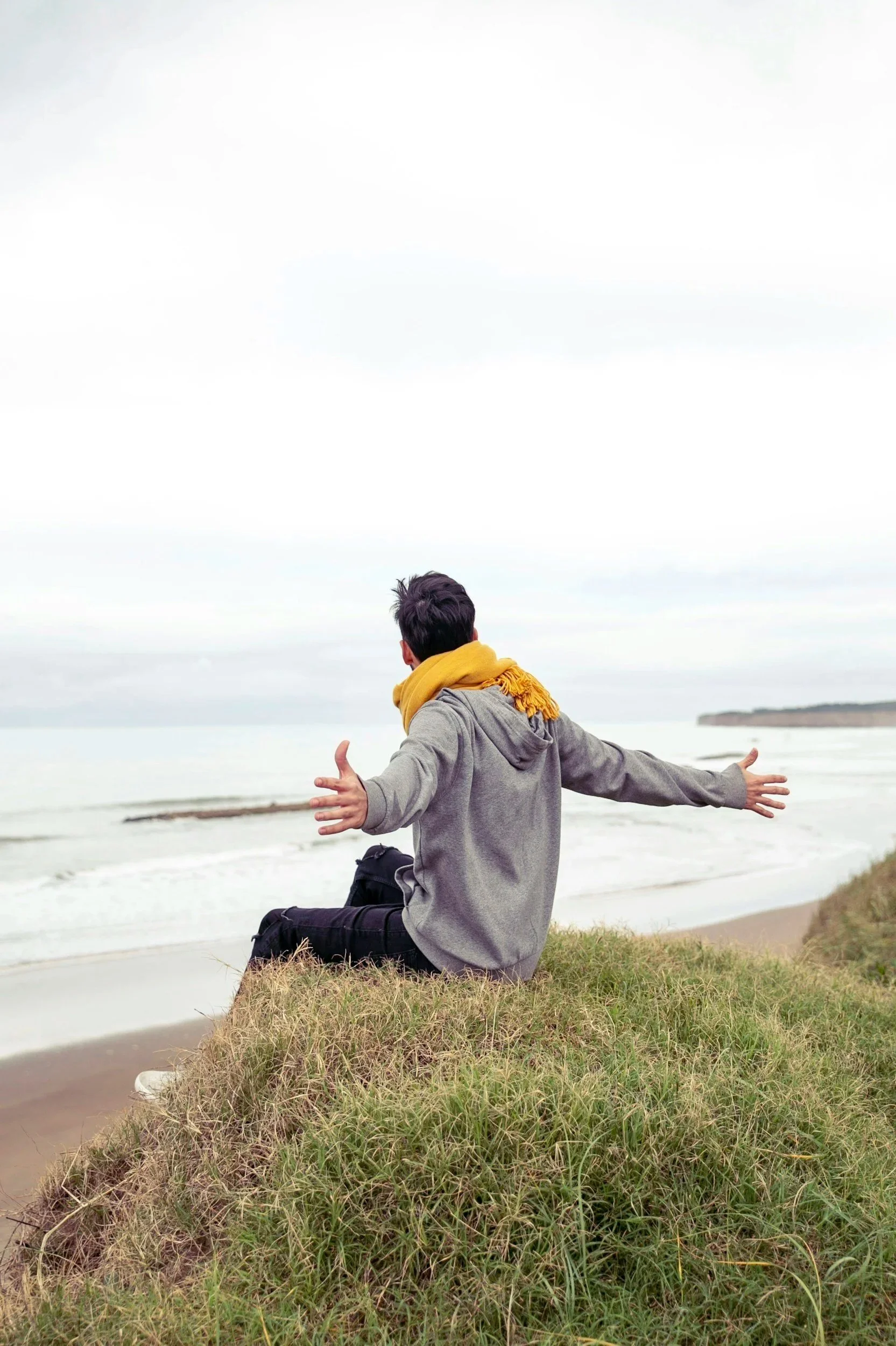 Person sitting on grass near a beach, wearing a grey hoodie and yellow scarf, with arms stretched out, facing the water under a cloudy sky.