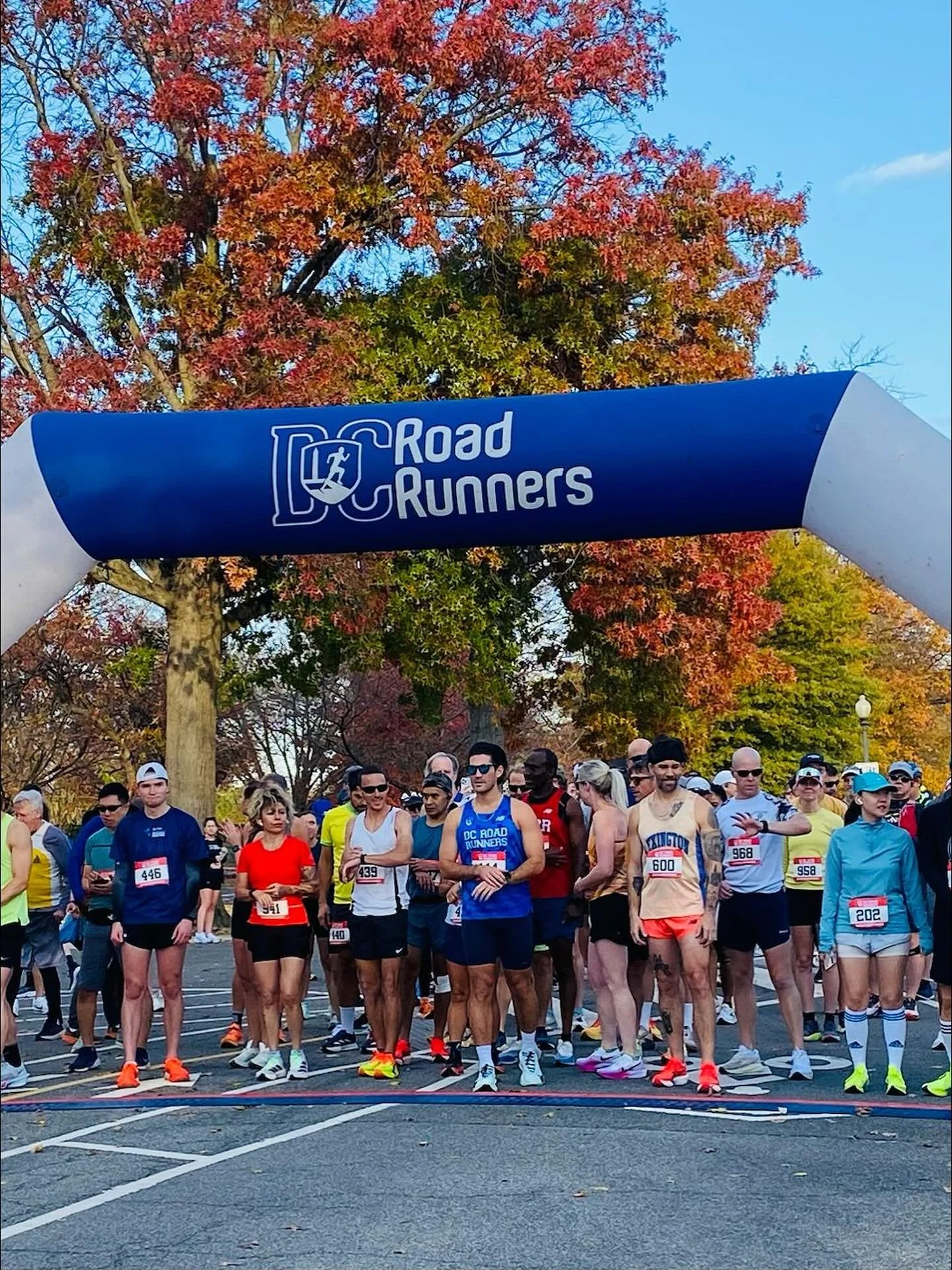 A diverse group of runners gathered at the starting line of a race, under a blue archway labeled 'DC Road Runners,' with trees displaying fall foliage in the background.