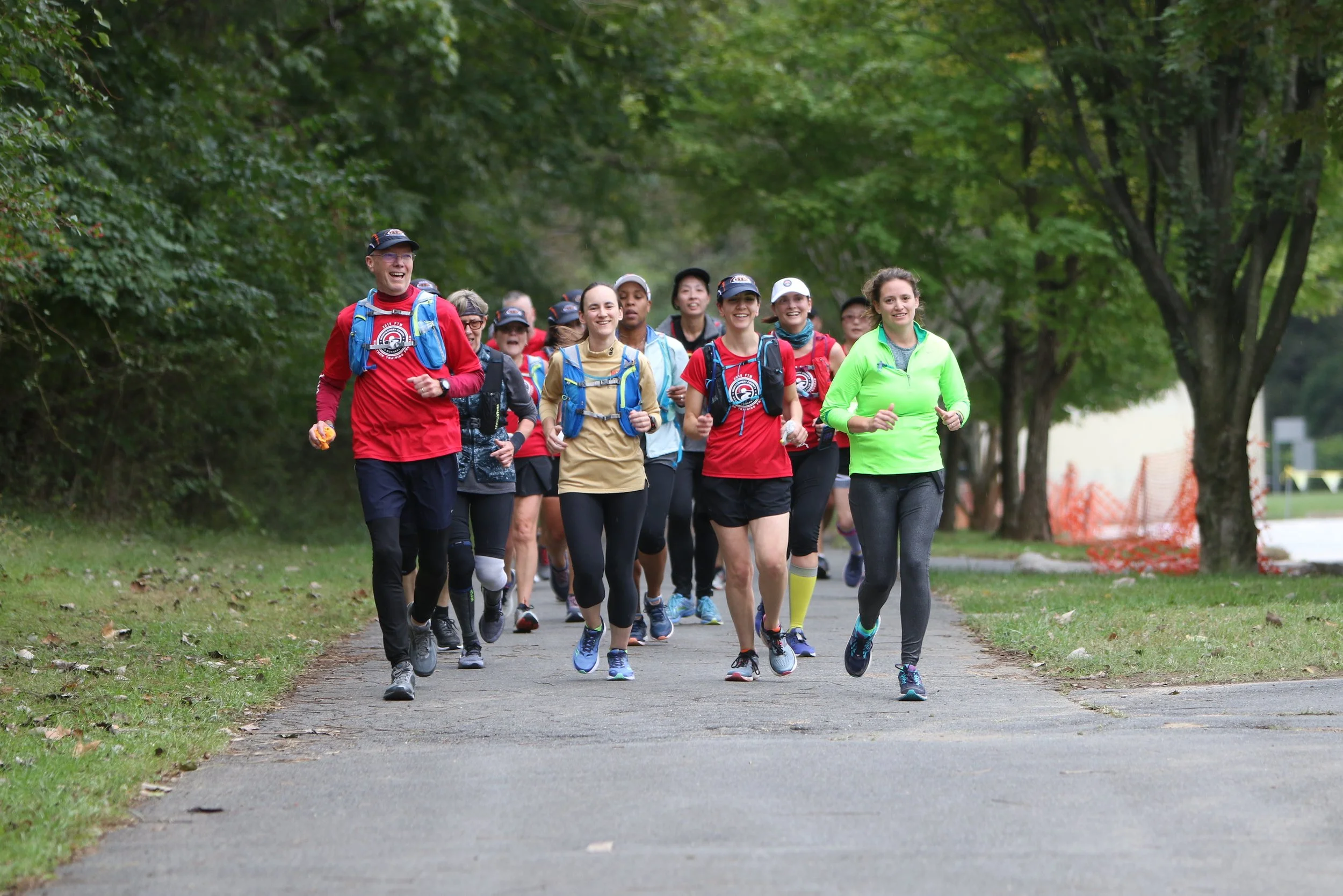 Group of people running outdoors on a paved path, surrounded by green trees, during daytime.