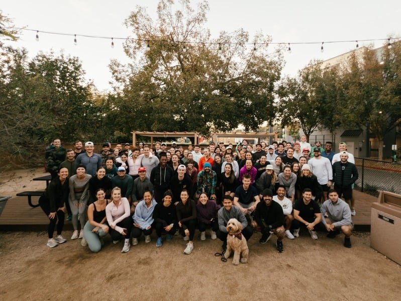A large group of people gathered outdoors for a group photo, with some sitting in front and others standing behind. There are trees and string lights overhead, and a dog sitting in front of the group.