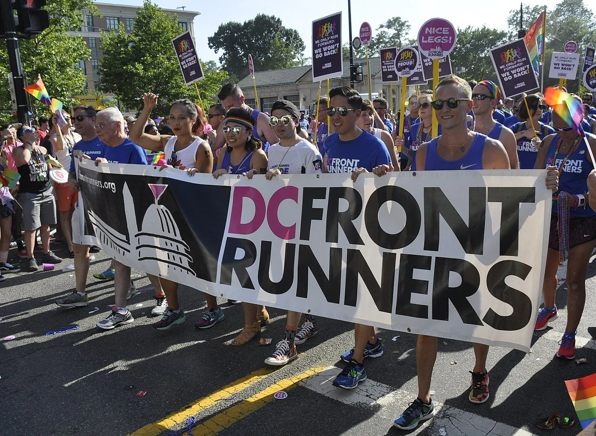 Group of people participating in a pride parade holding a large banner that reads "DC Front Runners" with a logo of the U.S. Capitol building, rainbow flags, and signs supporting LGBTQ+ rights.