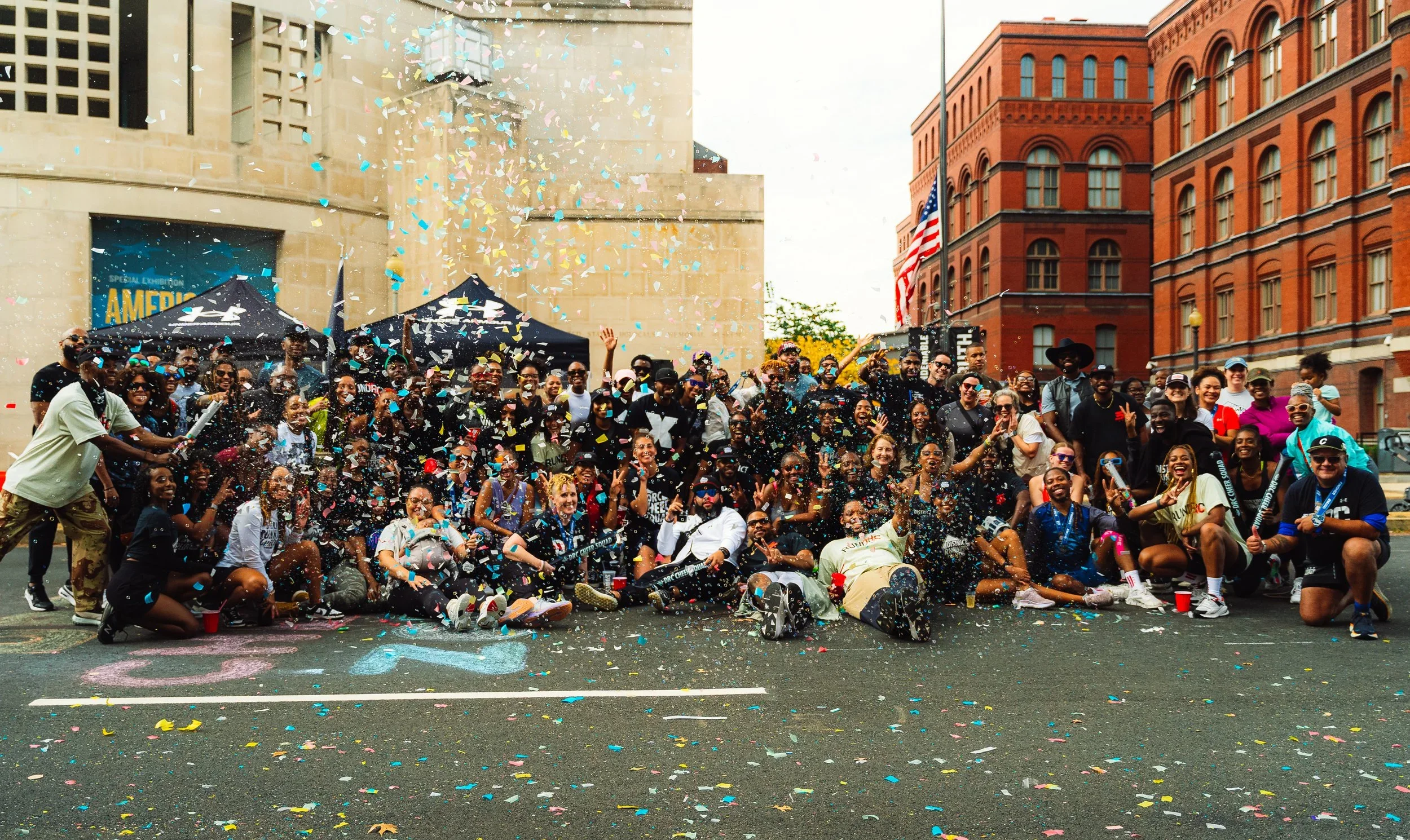 Group of people celebrating with confetti on an urban street, with tents and historic brick buildings in the background.