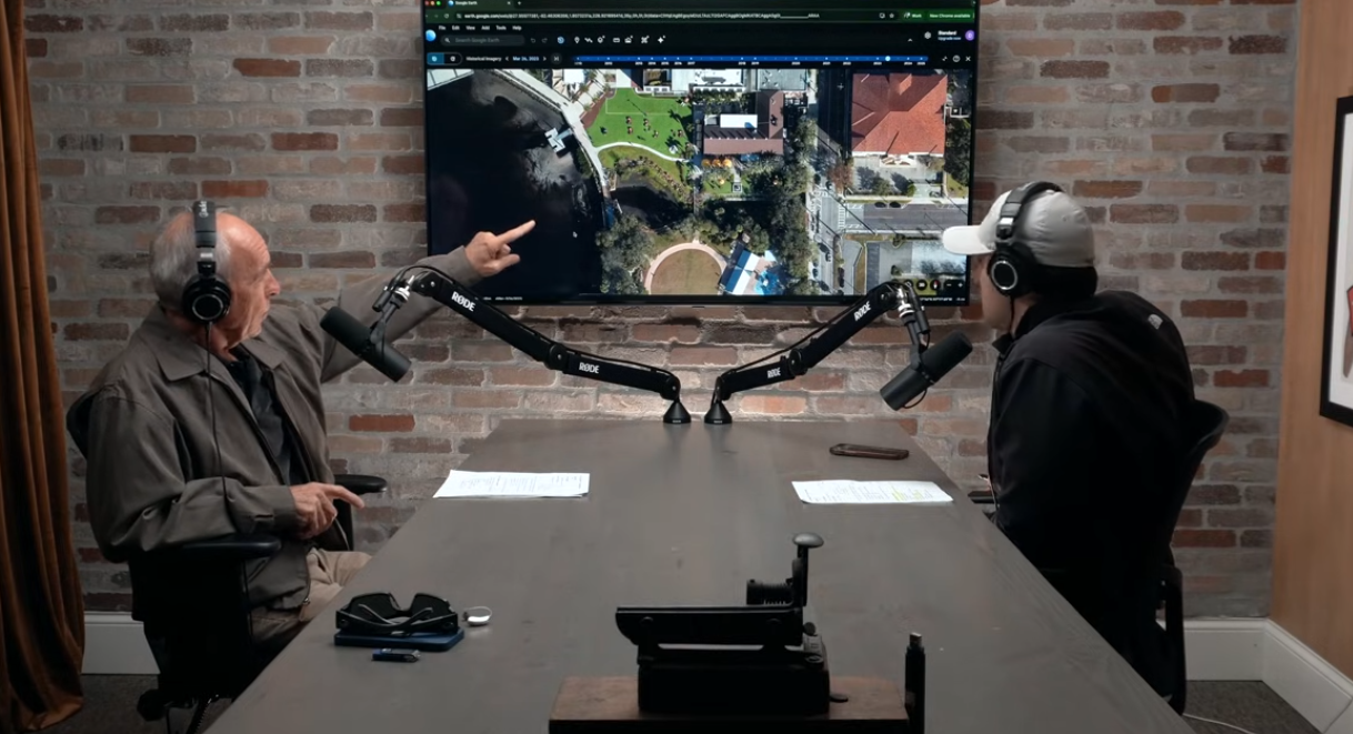 Two men are seated at a table in a podcast studio with a brick wall background and a large screen displaying an aerial view of a park and surrounding buildings. They are wearing headphones and speaking into microphones.