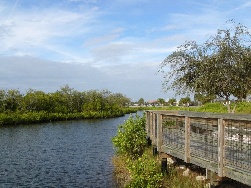 A wooden walkway with a metal railing runs along a small waterway, surrounded by green bushes and trees, with a partly cloudy sky overhead.