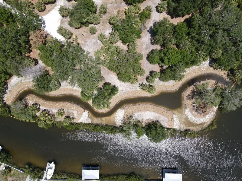 Aerial view of a winding creek surrounded by trees and a riverside area with some boats and structures.