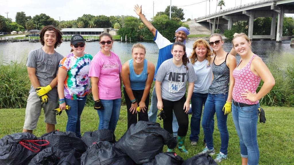 Group of nine diverse people smiling outdoors near a body of water, holding trash bags, posing after a cleanup event.