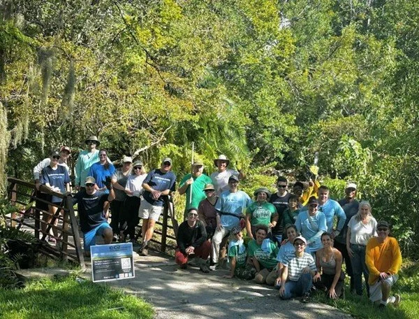 Group of people posing for a photo outdoors on a sunny day, with green trees and a small wooden bridge in the background.