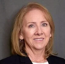 Portrait of a middle-aged woman with blonde hair, wearing a dark blazer, smiling slightly at the camera.