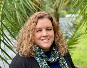 A woman with curly blonde hair smiling outdoors with green palm leaves in the background.