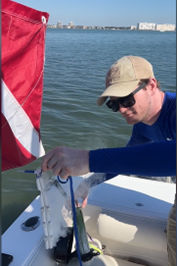 A man wearing sunglasses and a beige cap is on a boat holding a fish he caught, with water and a distant city skyline in the background.