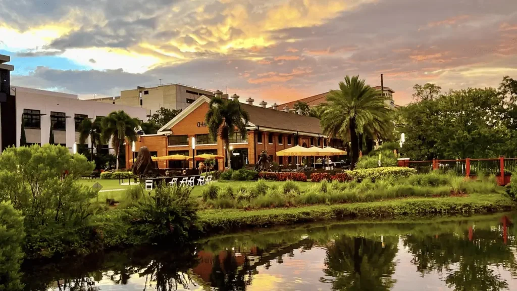A scenic view of a restaurant or cafe with outdoor seating, surrounded by lush greenery and palm trees, at sunset, with a pond in the foreground reflecting the sky and trees.