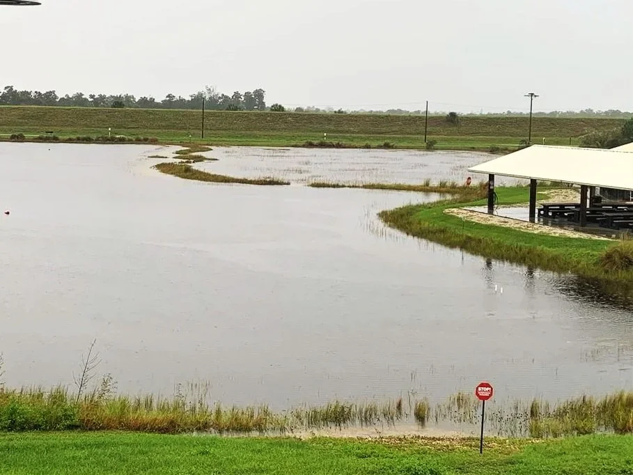 A large body of water, possibly a flooded area, with grassy patches and a pavilion on the right side. The scene appears overcast with a cloudy sky.