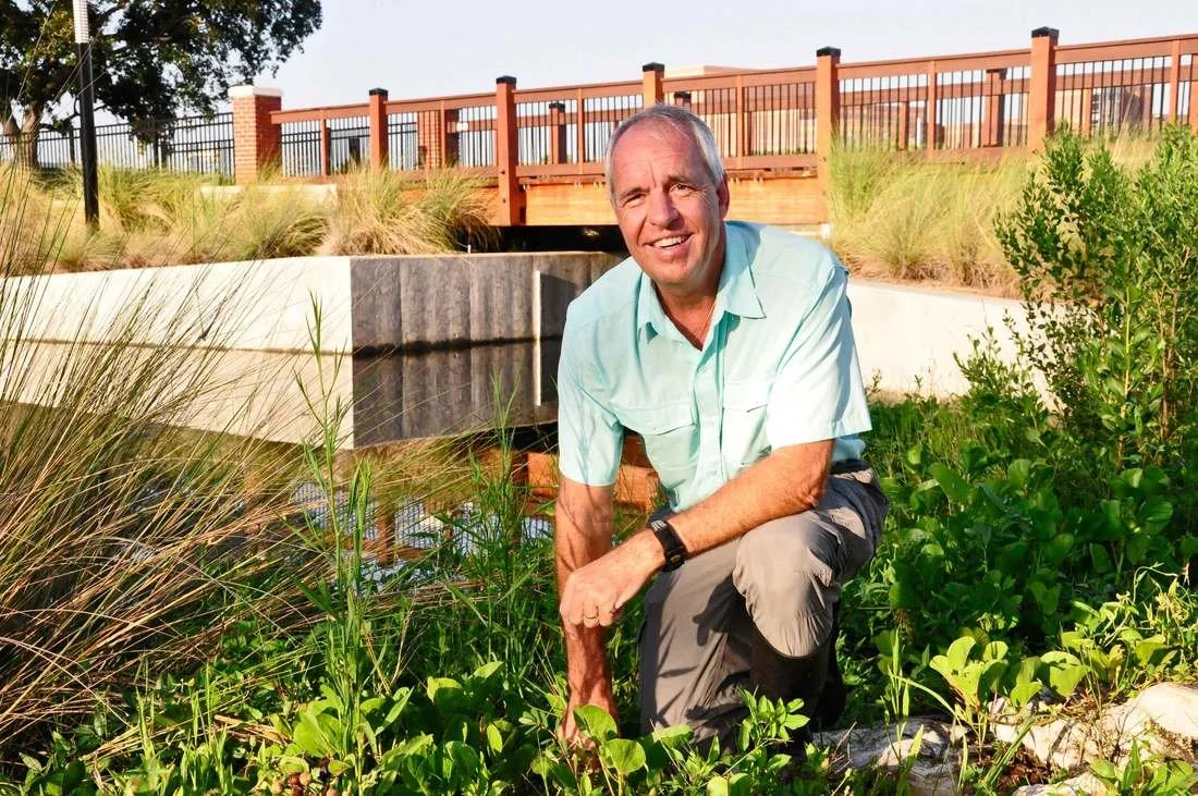 A man kneeling outdoors near green plants and tall grasses, with a wooden fence or bridge in the background, under a clear sky.