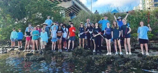 Group of young volunteers standing on rocks by water during daytime, some holding paddles, outdoors in an urban park.