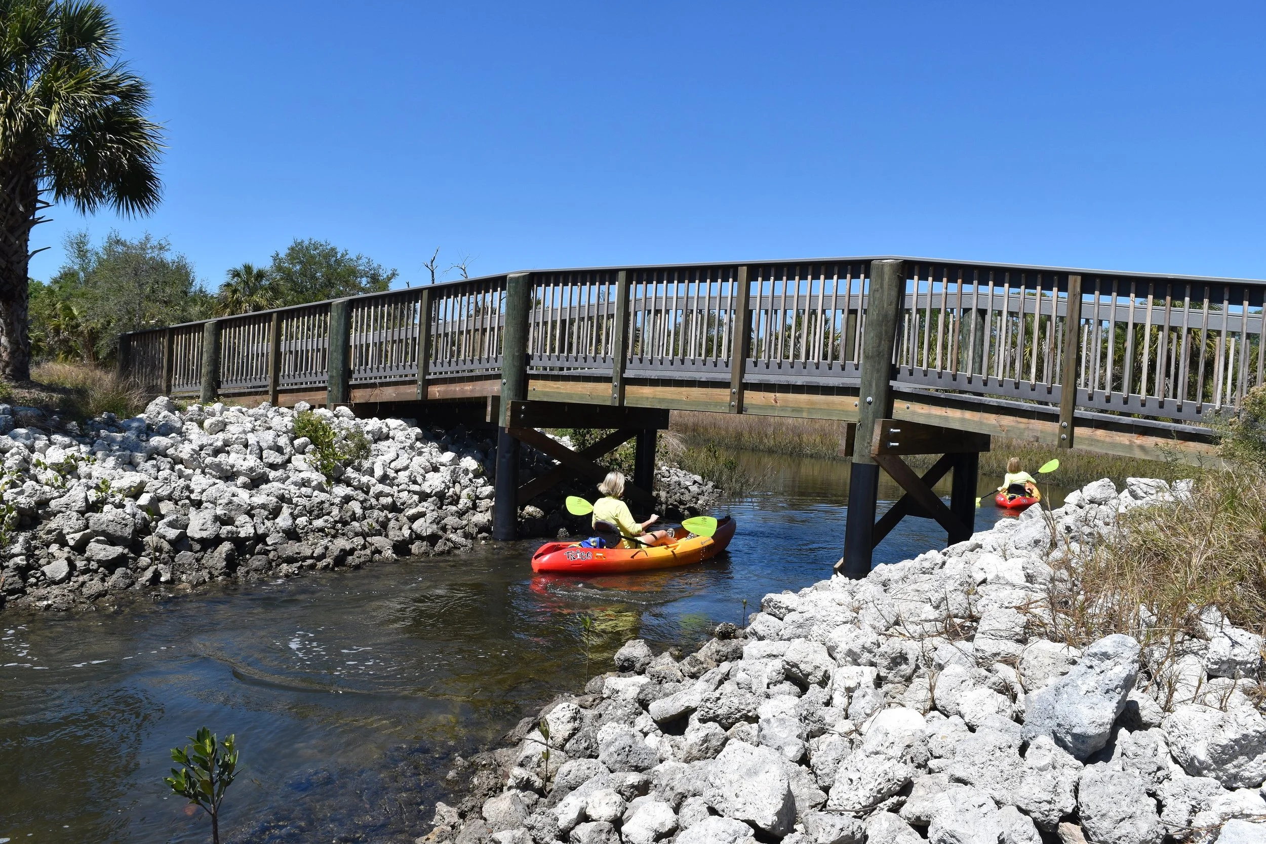 Two children in yellow life jackets kayaking under a wooden bridge over a small creek surrounded by rocks and greenery on a sunny day.