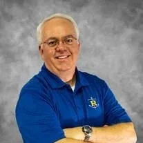 Portrait of a smiling man with glasses, wearing a blue polo shirt, arms crossed, against a gray background.