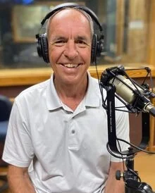 A smiling man wearing headphones in a recording studio with a microphone in front of him.