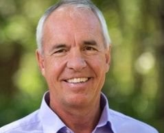 Tom smiling, wearing a light blue collared shirt outdoors with blurred green foliage in the background.