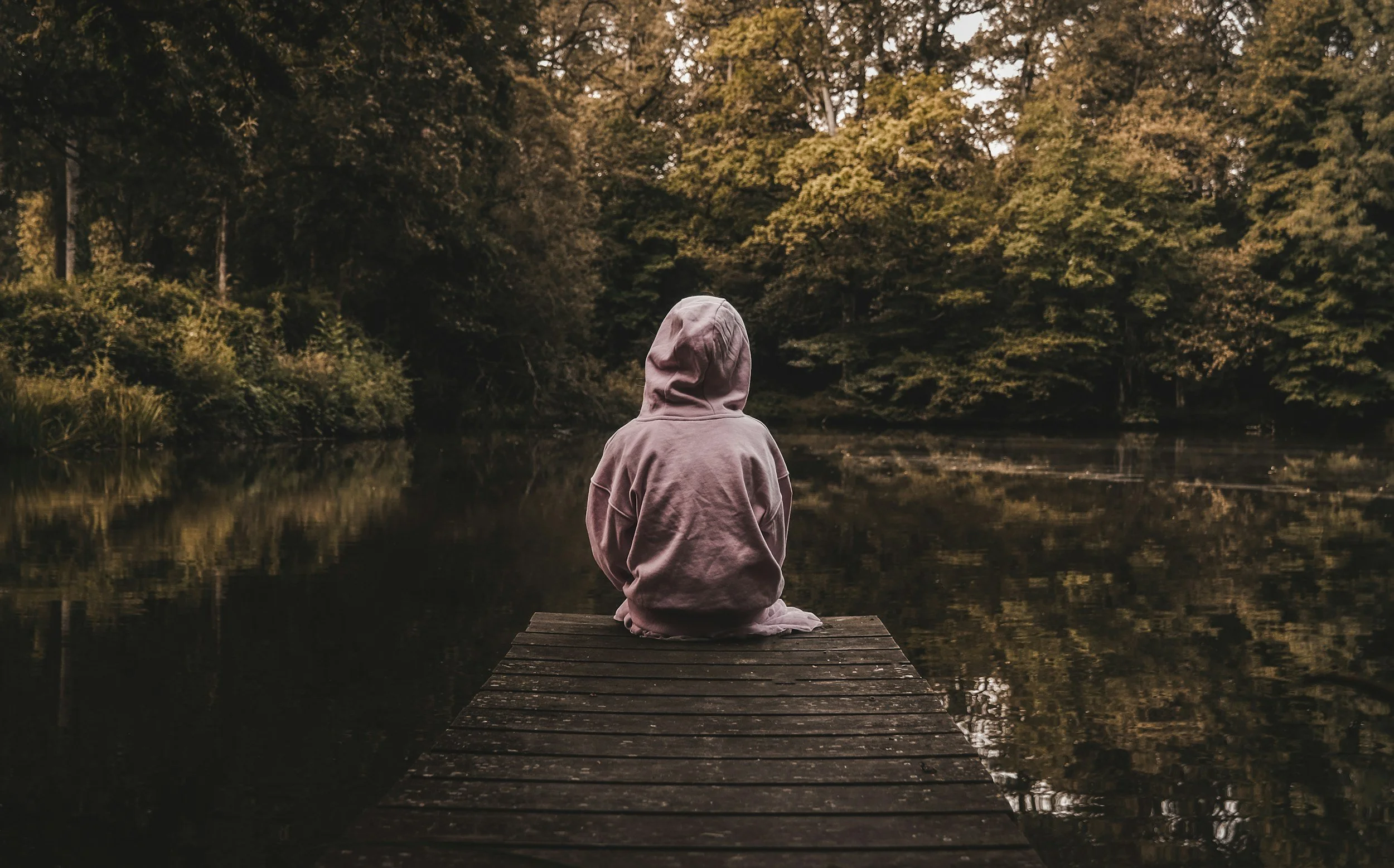 A person in a hoodie sitting on a dock by a calm lake, facing away, surrounded by trees during autumn.
