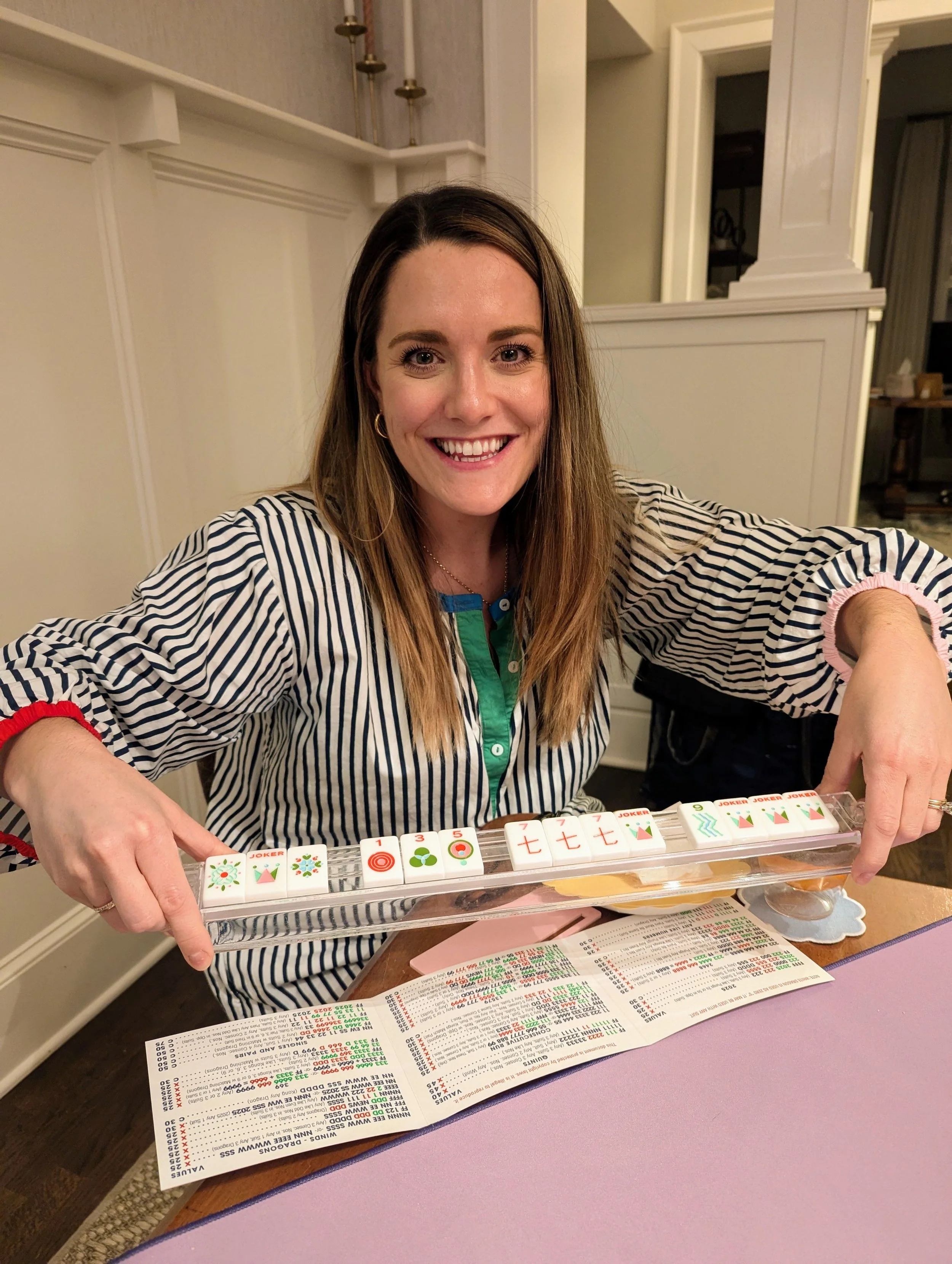 Woman smiling and playing Mahjong, with Mahjong tiles on the table and a game score sheet.