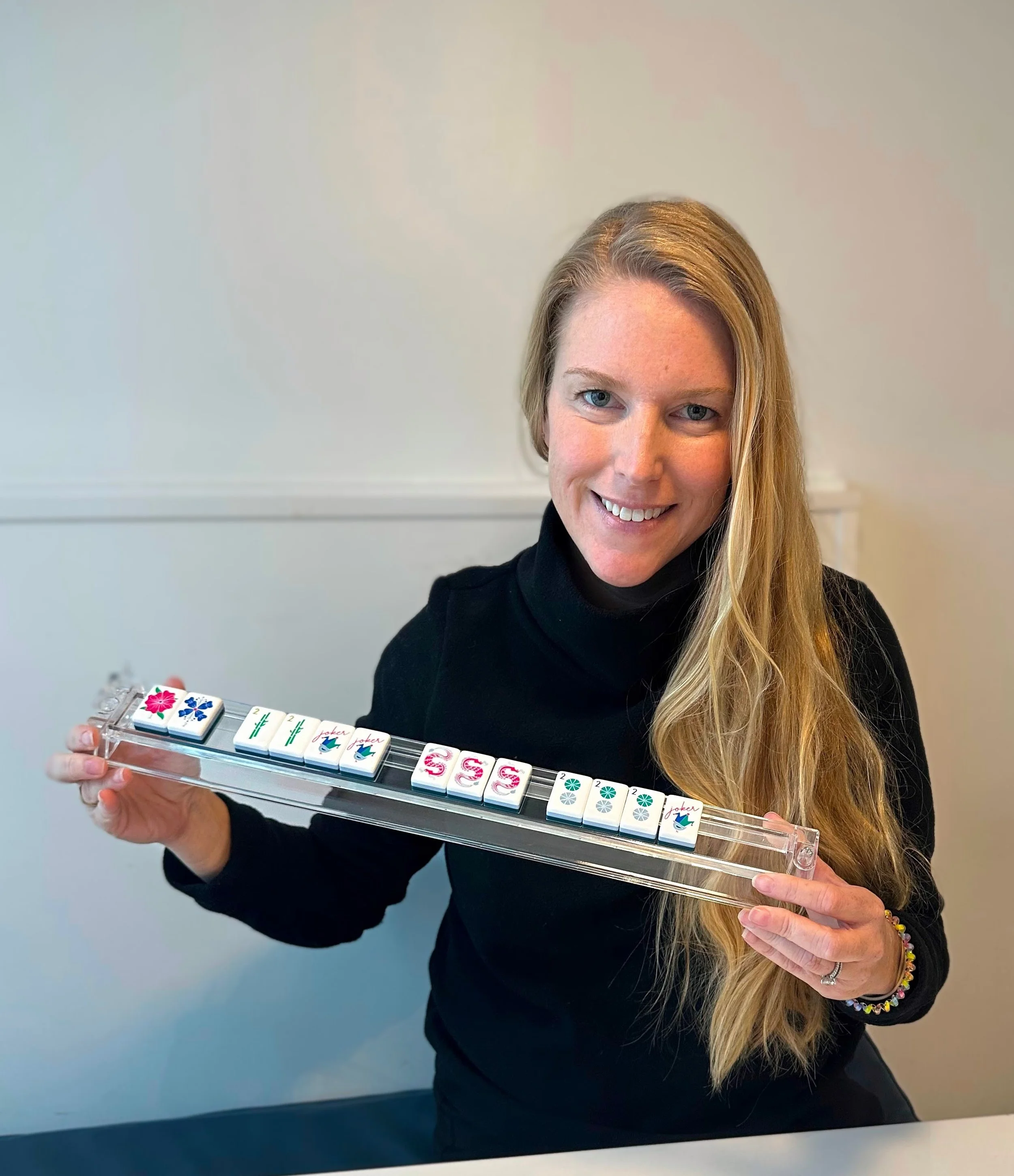Woman with long red hair smiling and holding a mahjong tile rack with various tiles.