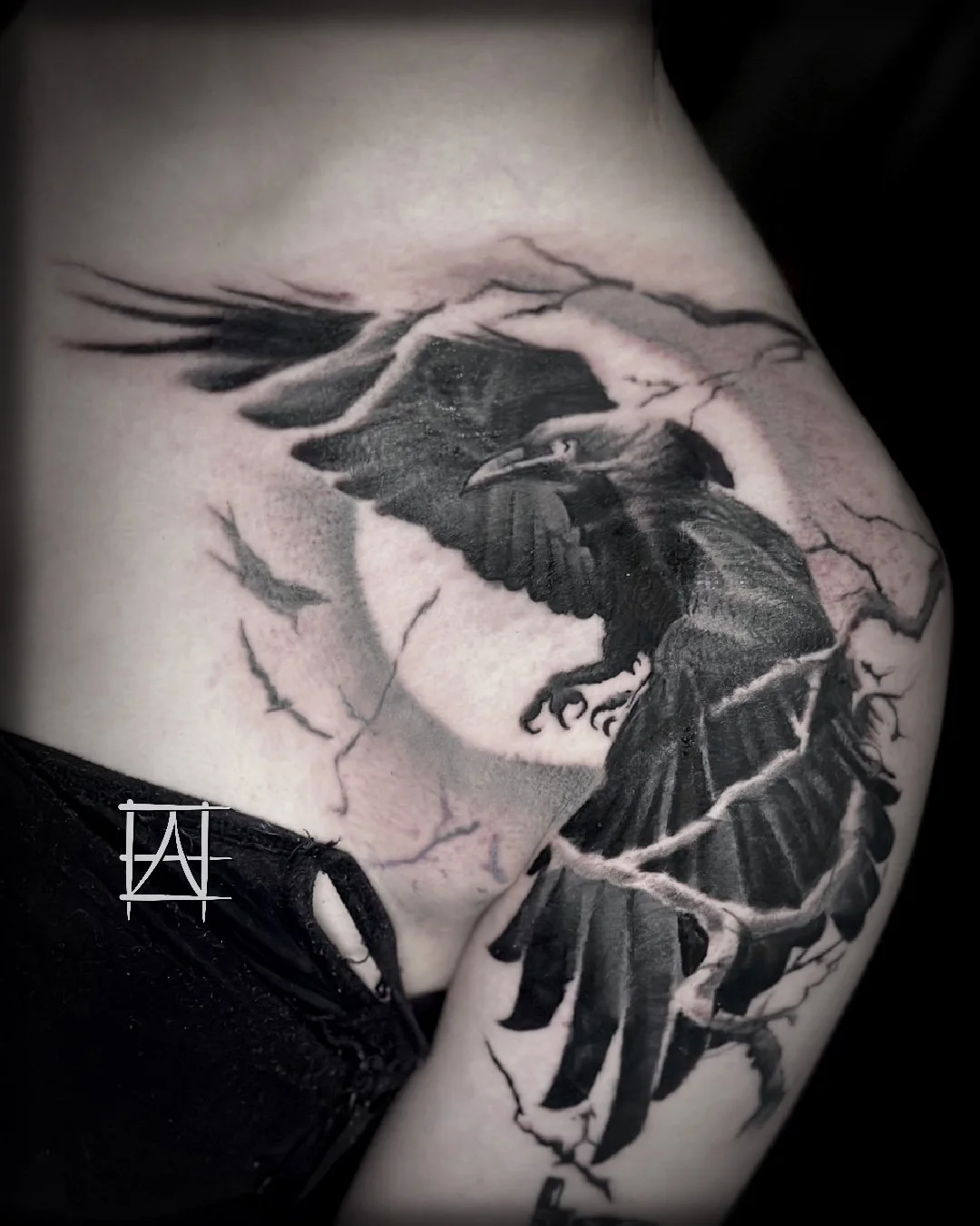 A black and gray tattoo of a raven with wings spread, perched on a branch with cracks, on someone's skin.