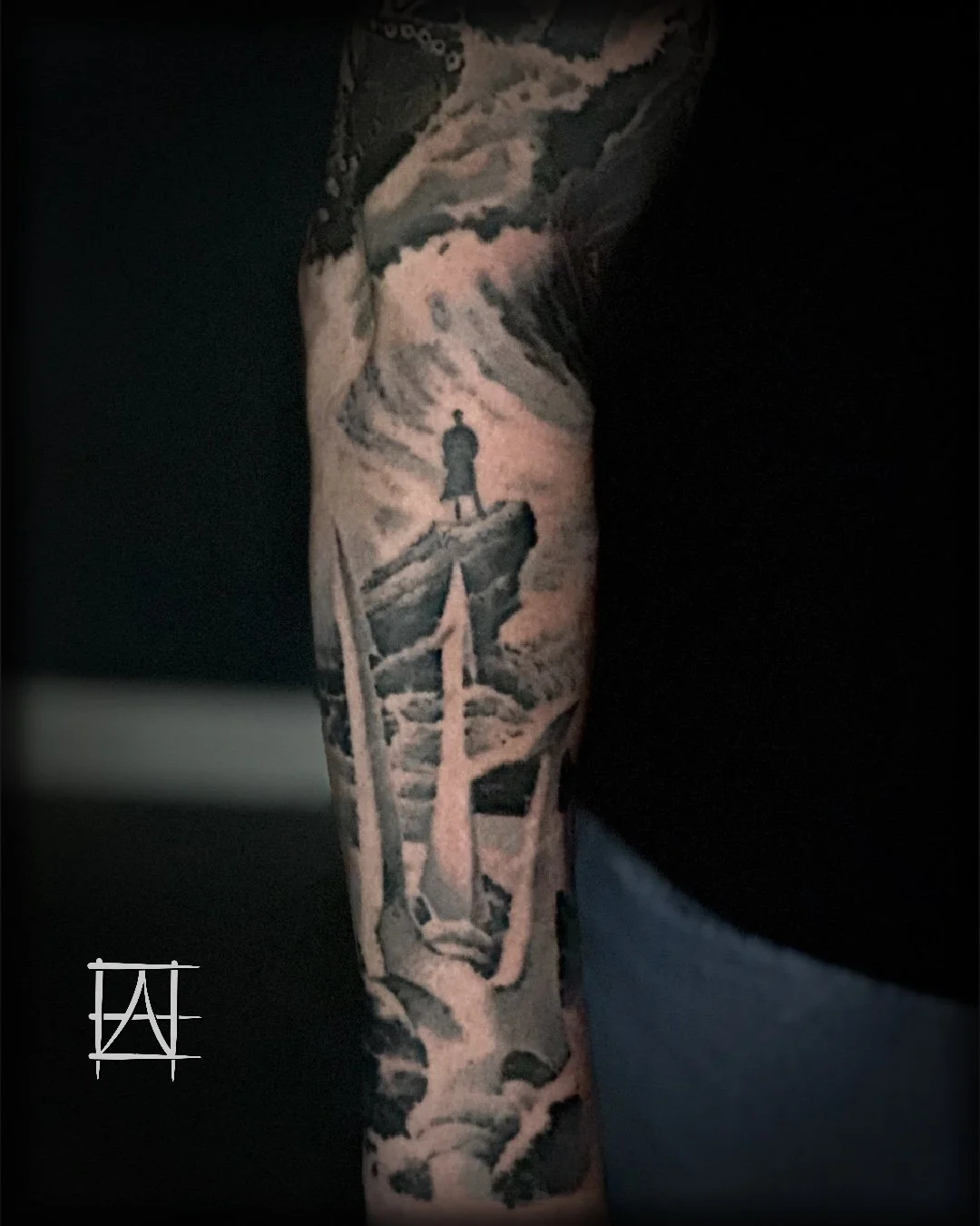 Black and gray tattoo of a person standing on a rocky ledge, overlooking a mountainous landscape.