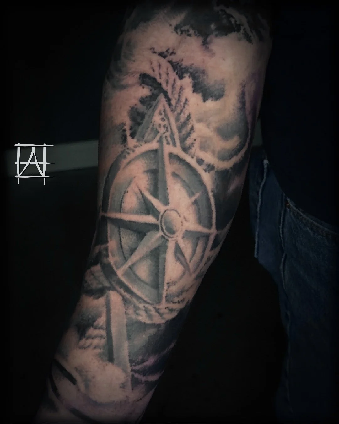Black and grey tattoo of a compass with a star, surrounded by clouds and a tree branch on a person's arm.