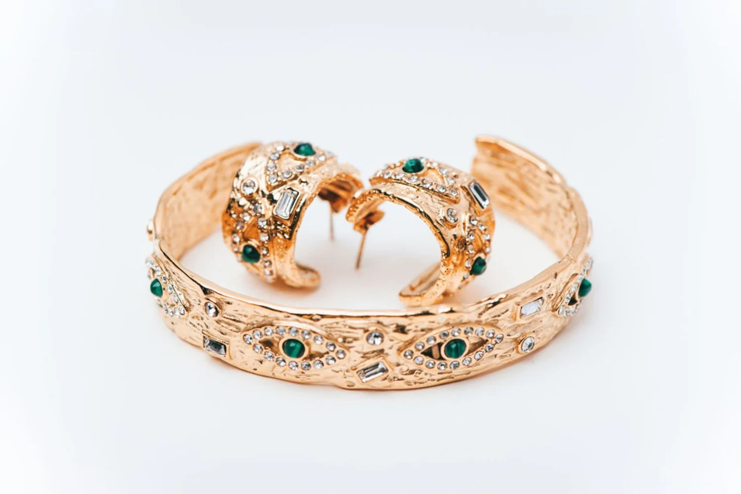 Gold jewelry set consisting of a bracelet and earrings decorated with green stones, clear rhinestones, and intricate designs, displayed on a white background.