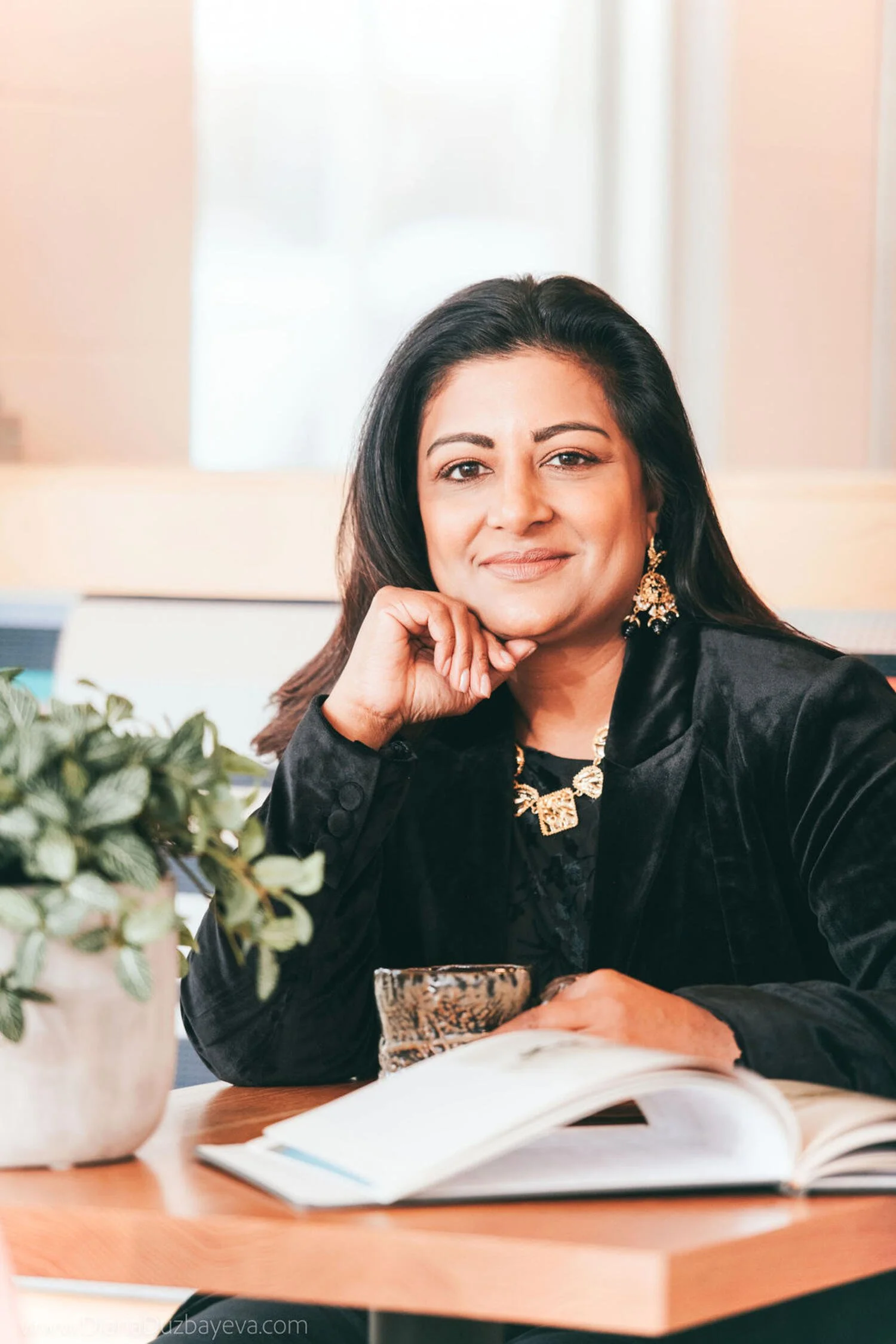 A woman with dark hair wearing gold jewelry, a black velvet blazer, and earrings, sitting at a wooden table with an open book, a plant, and a cup, smiling at the camera in an indoor setting.