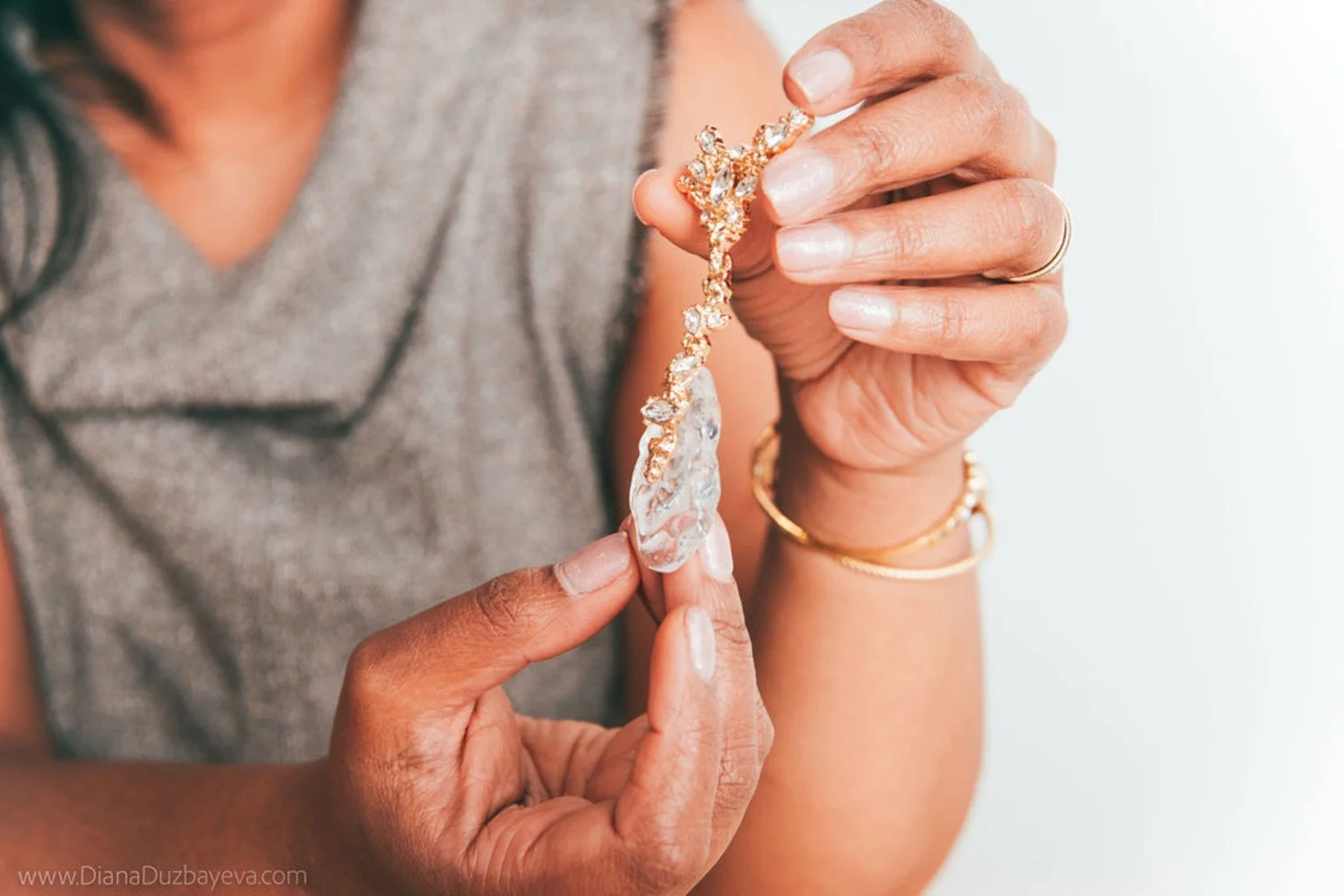 Close-up of a person holding a large, ornate gold necklace with sparkling diamonds, adjusting it with their fingers. The person wears gold bracelets and a gray sleeveless garment.