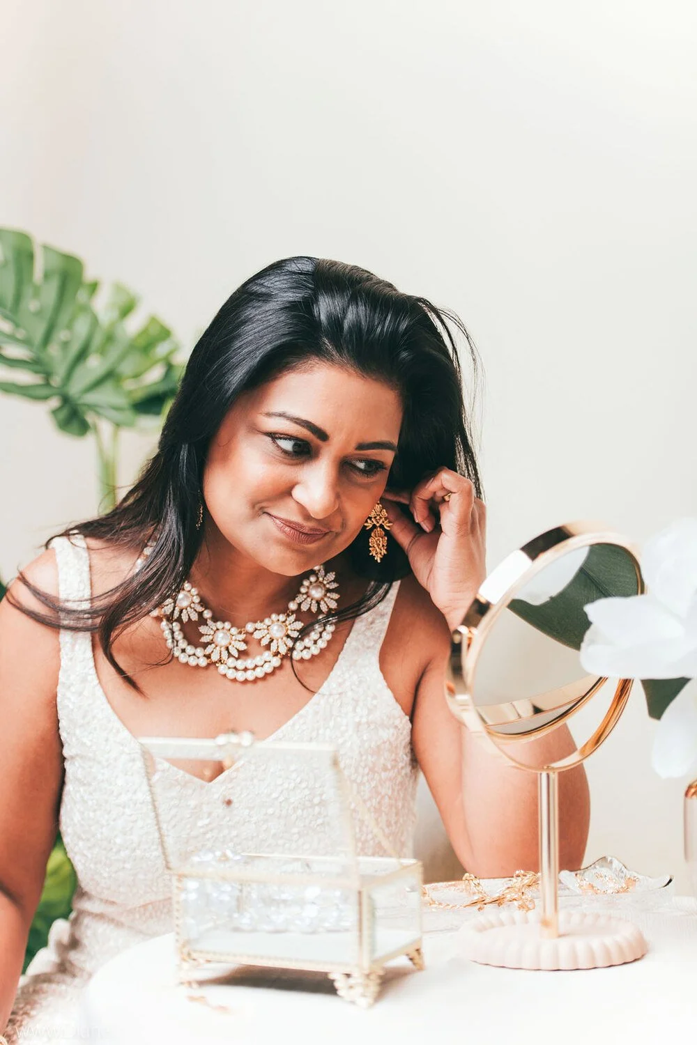 A woman looking in a hand mirror, wearing pearl jewelry and a white dress, with a large green plant in the background.