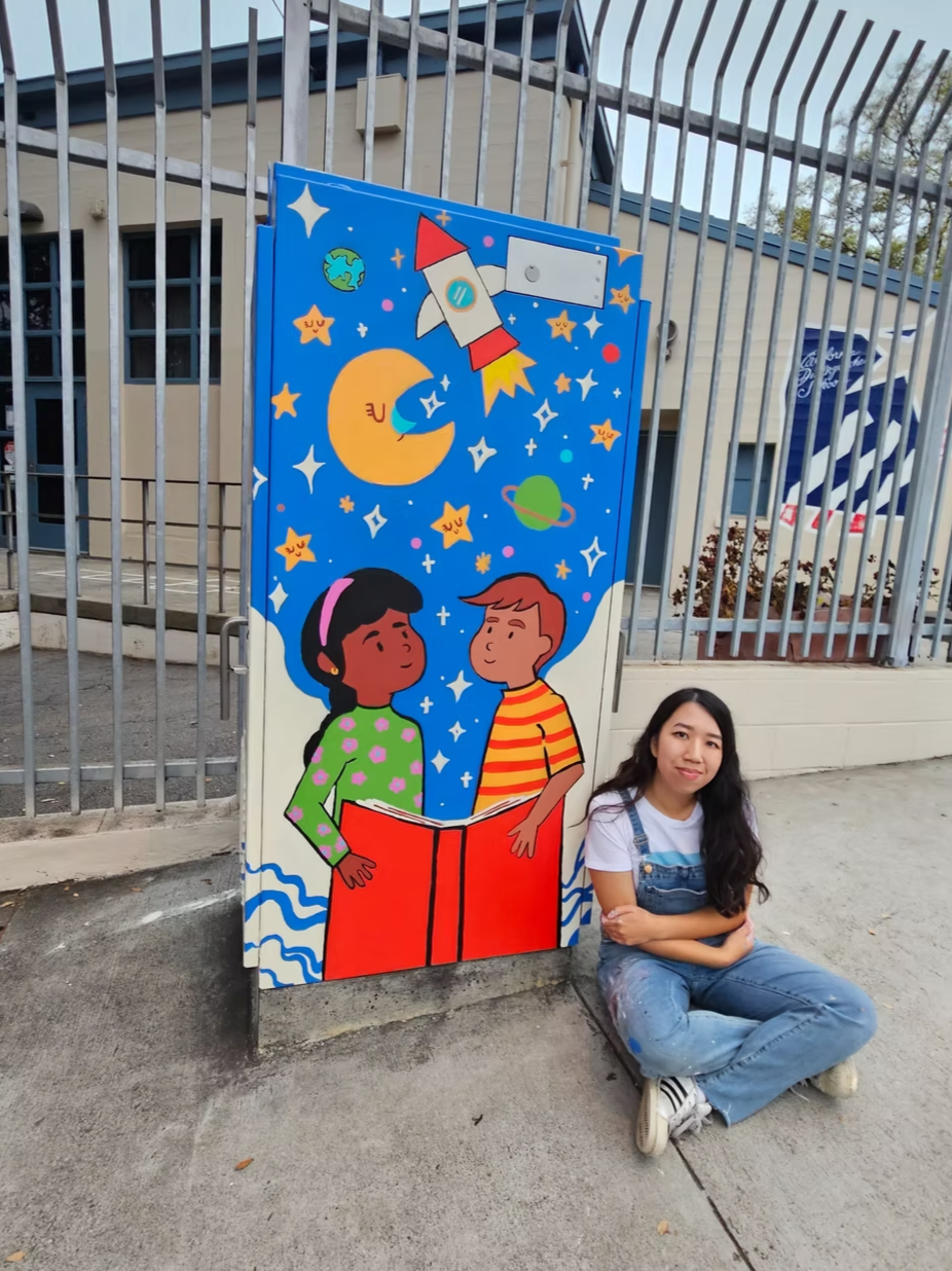 A woman sitting on the ground next to a colorful mural featuring two children reading a book, a rocket, stars, planets, and space elements.