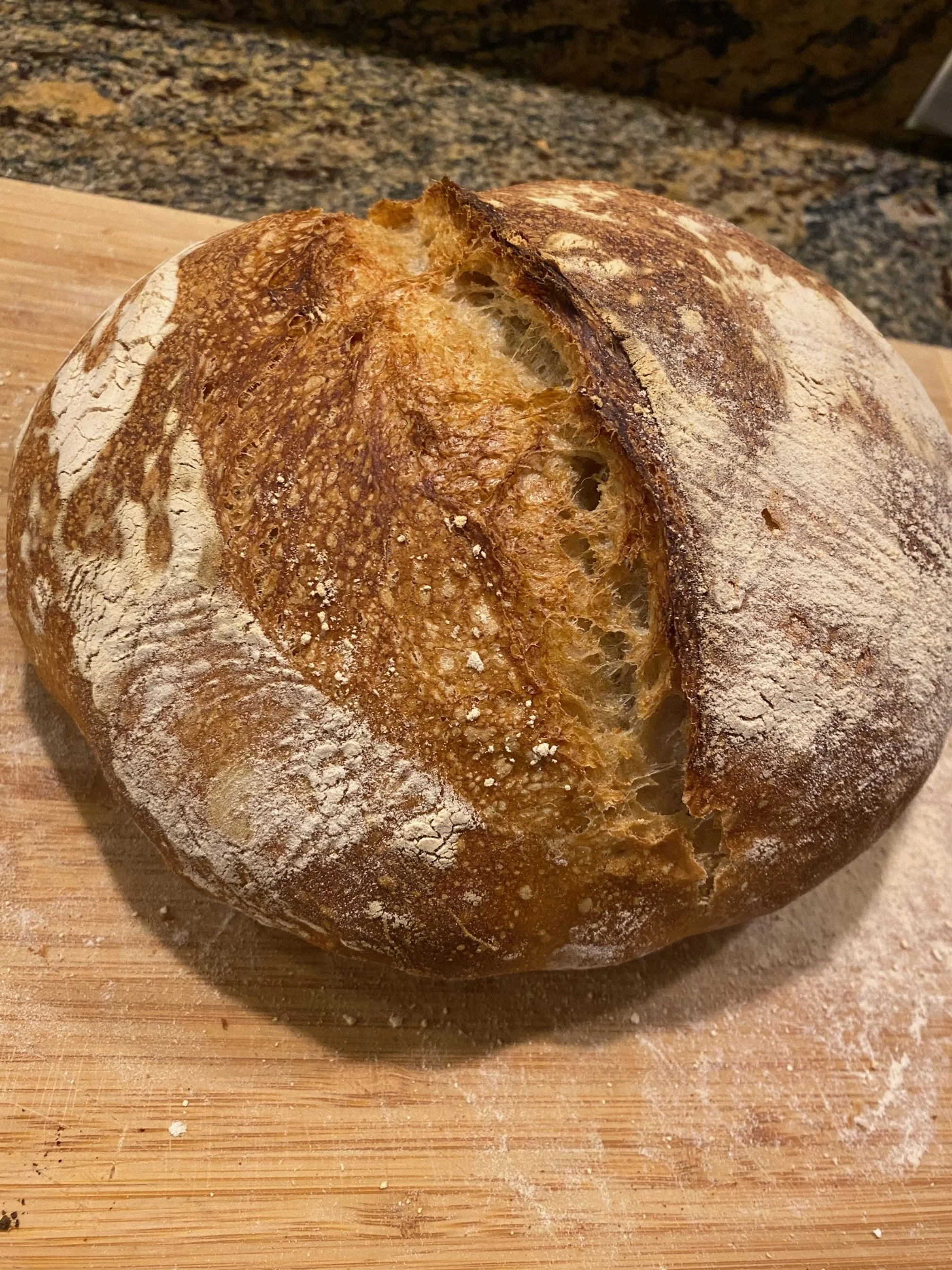 A round, rustic loaf of bread with a scored top crust, dusted with flour, resting on a wooden cutting board.