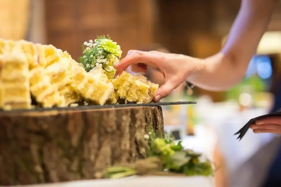 A person is serving or cutting a slice of layered cake on a table decorated with flowers, with the cake placed on a wooden surface.