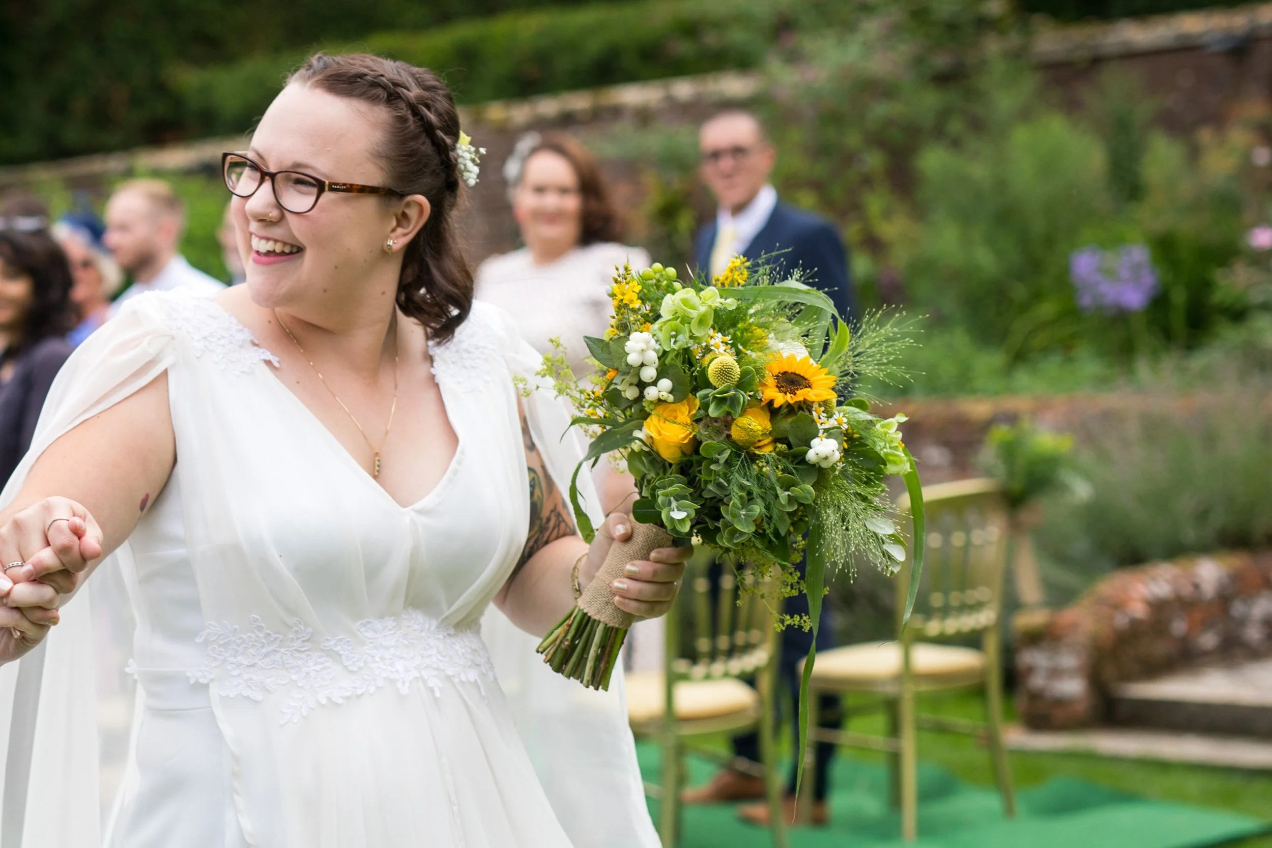 A woman in a white dress holding a vibrant yellow and green bouquet, smiling outdoors during a wedding ceremony.
