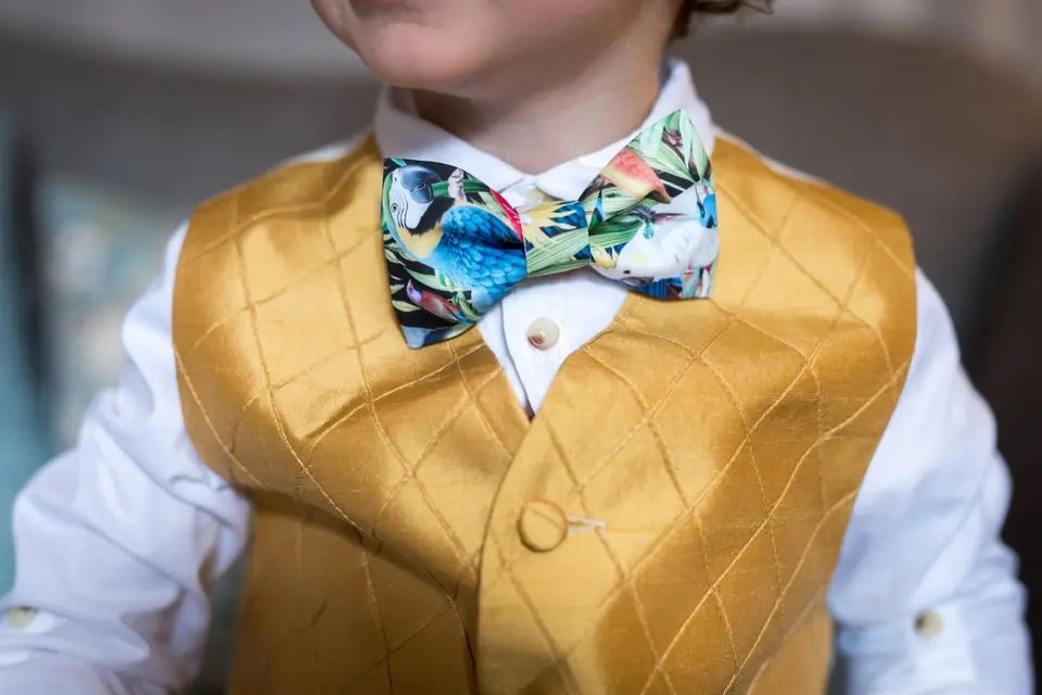Close-up of a young boy wearing a white dress shirt, a yellow vest with a diamond pattern, and a colorful tropical bird print bow tie.