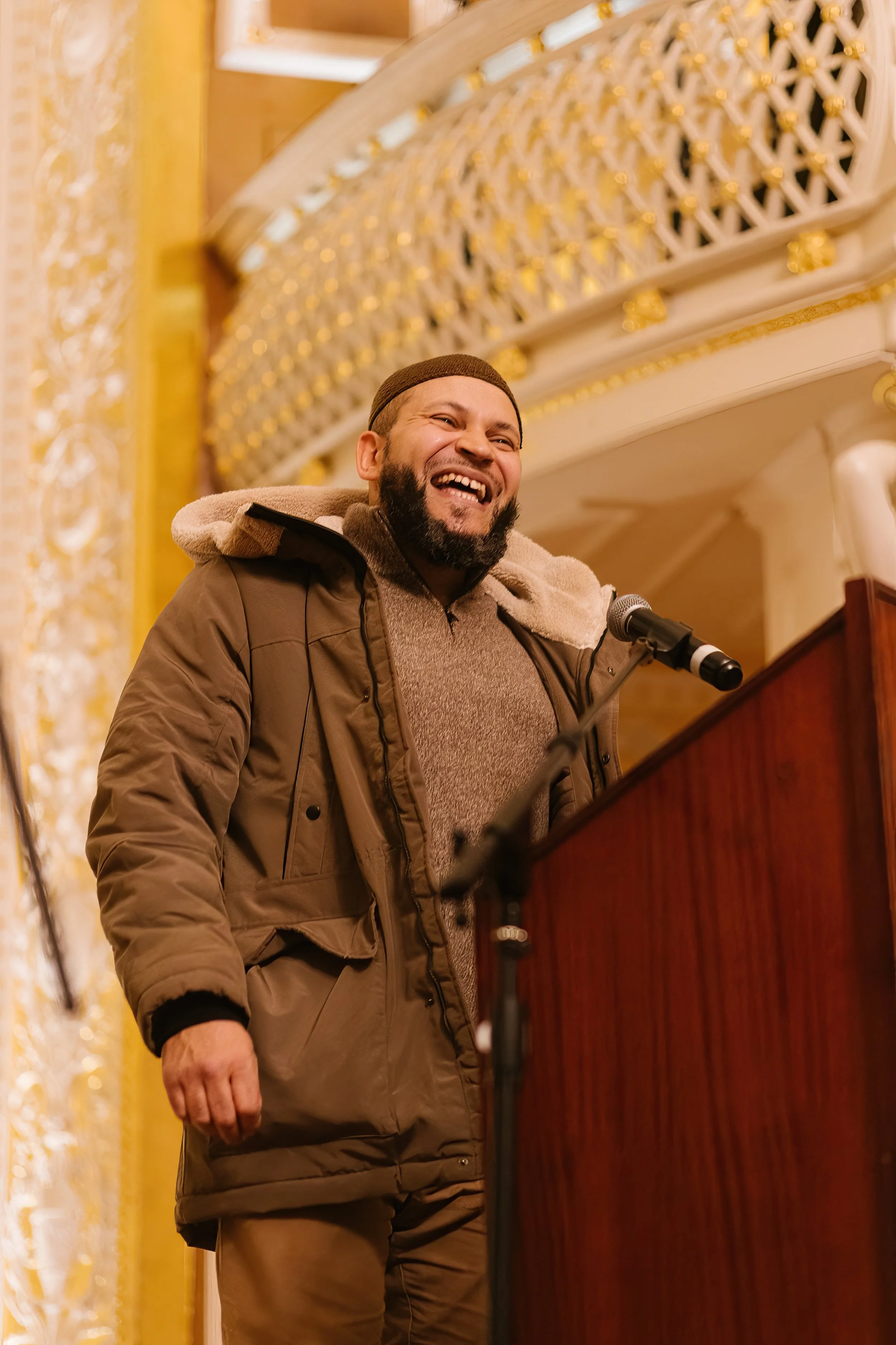 A man with a beard smiling and speaking at a podium in an ornate, gold-decorated room.
