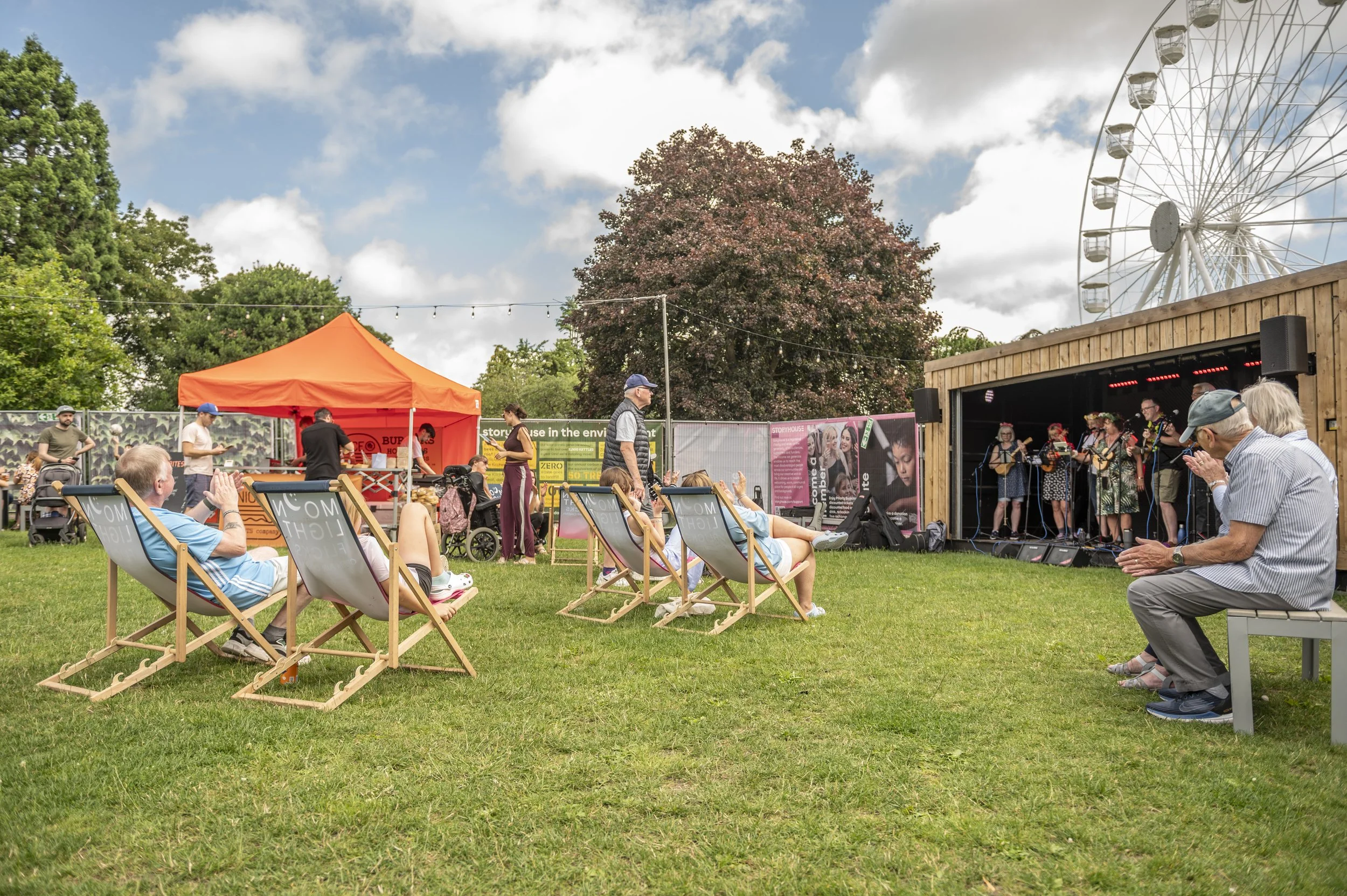 Ukuele band plays in an outdoor container in front of a fressis wheel and an audience