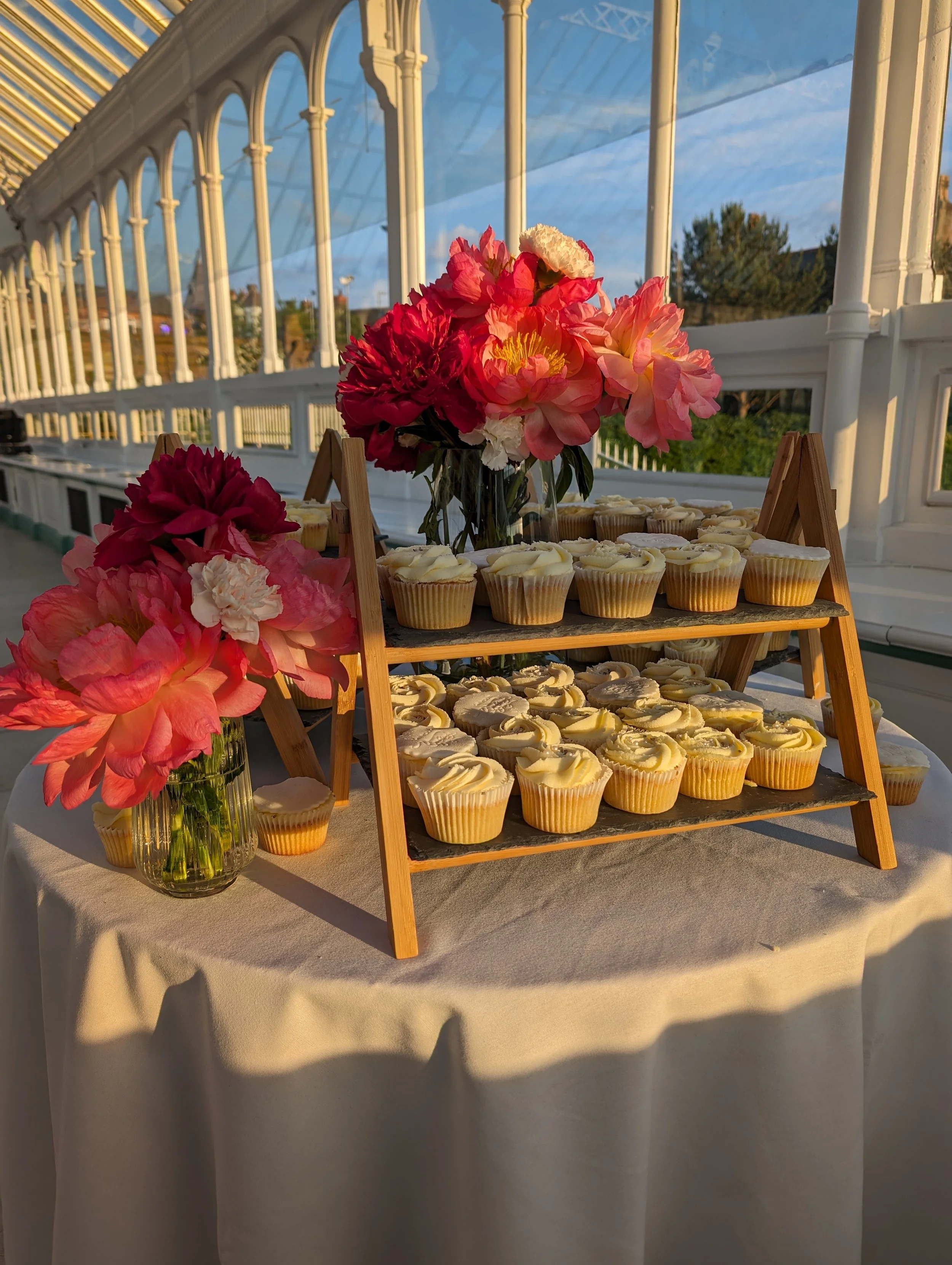 Cupcakes arranged on a wooden cupcake stand with pink and white flowers in a glass vase, set on a table with a white tablecloth in a sunlit greenhouse or pavilion.