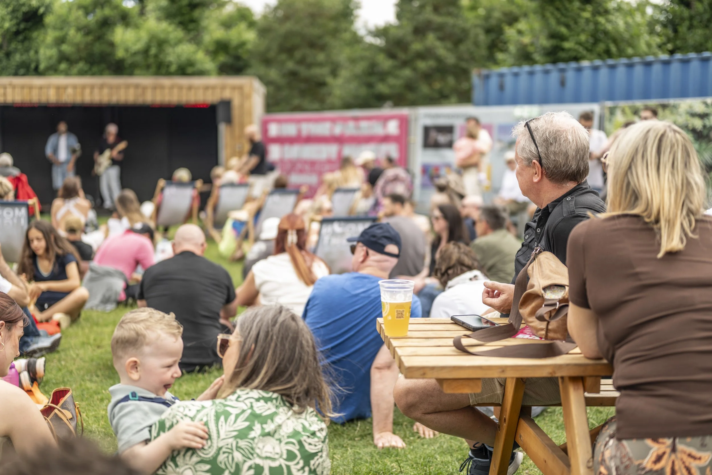 People sitting on grass and watching a live outdoor concert with a small stage and band playing, trees in the background, some with drinks, children and adults enjoying the event.