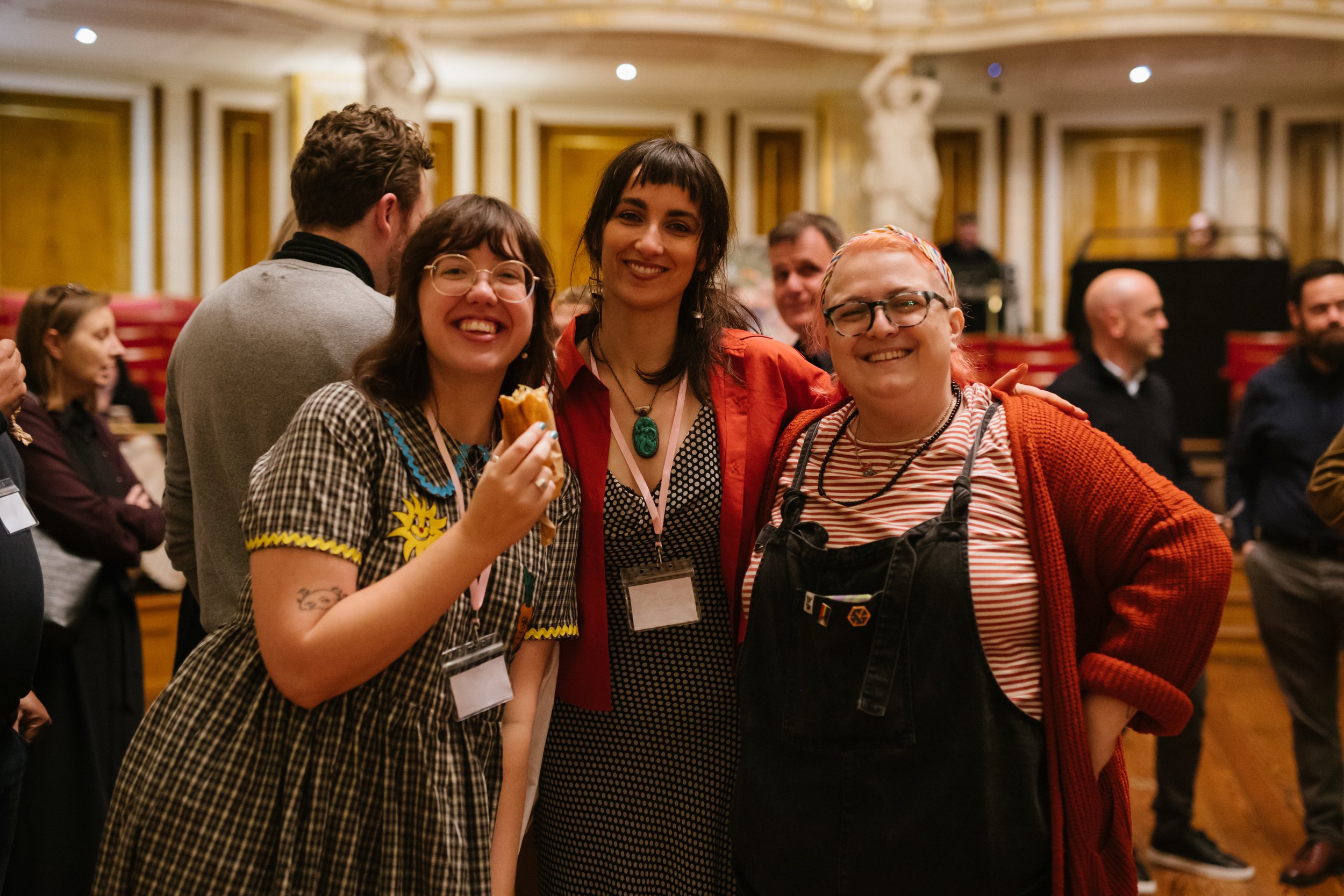 Three smiling women standing together at an indoor event. The woman on the left is wearing a checked dress with colorful embroidery, glasses, and is holding a snack. The woman in the middle has dark hair, is wearing a red jacket over a black and white dress, and a large green pendant necklace. The woman on the right has glasses, short reddish hair, and is wearing a striped shirt under black overalls with a red cardigan. There are other people and ornate decor in the background.