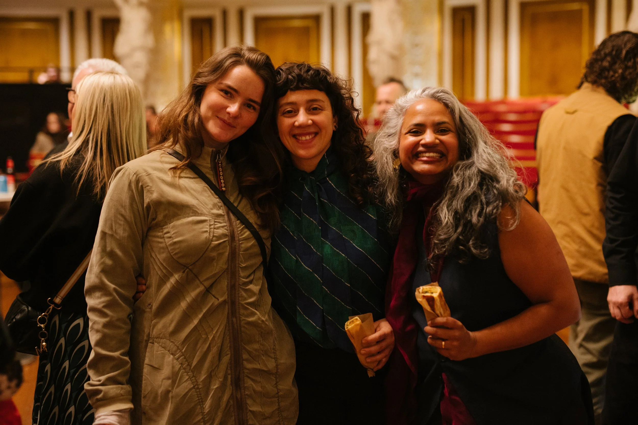 Three women smiling and standing close together, each holding a pastry, at an indoor gathering or celebration.