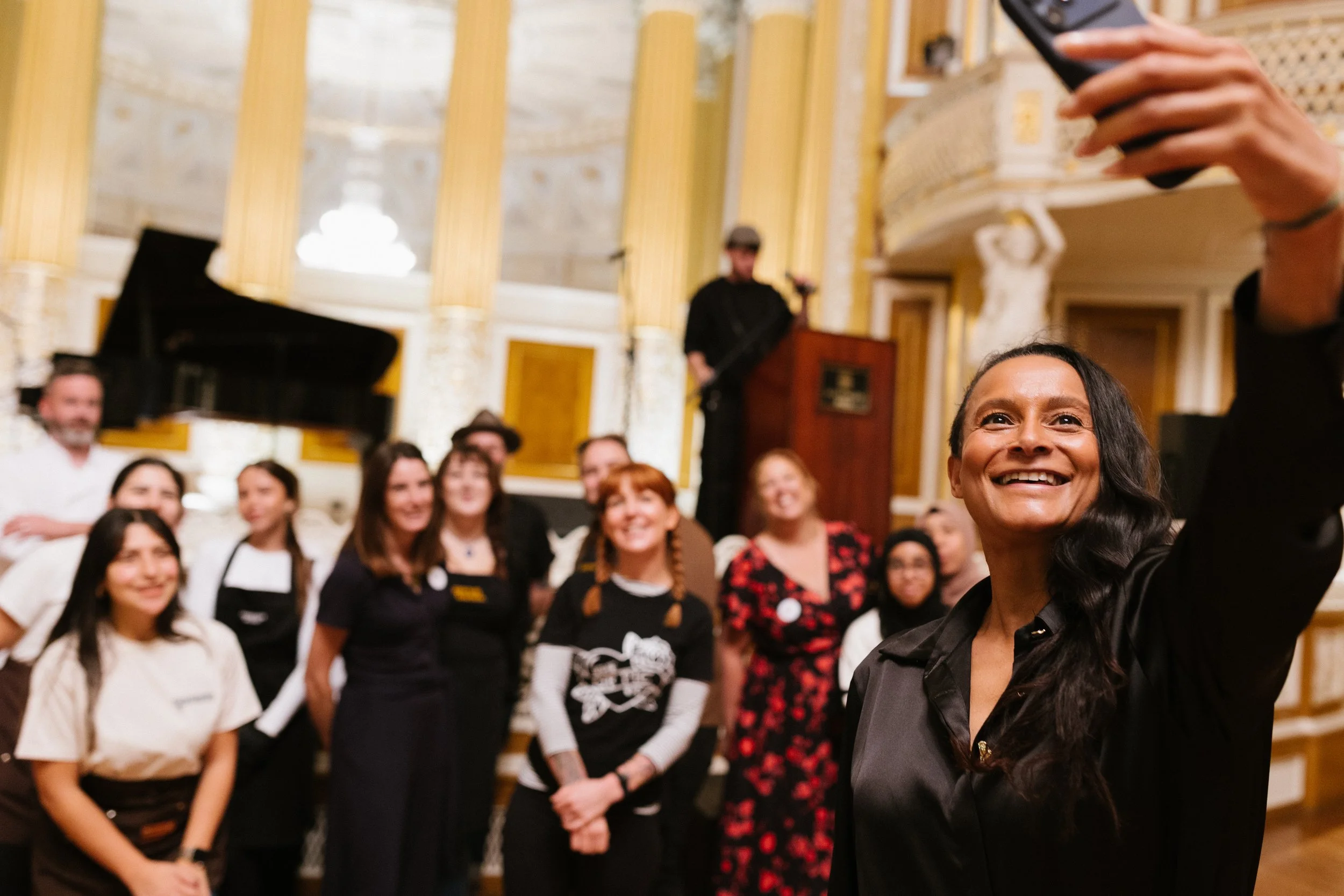 A woman takes a selfie at a social event or gathering, with a group of smiling people in the background inside a decorated hall or ballroom.