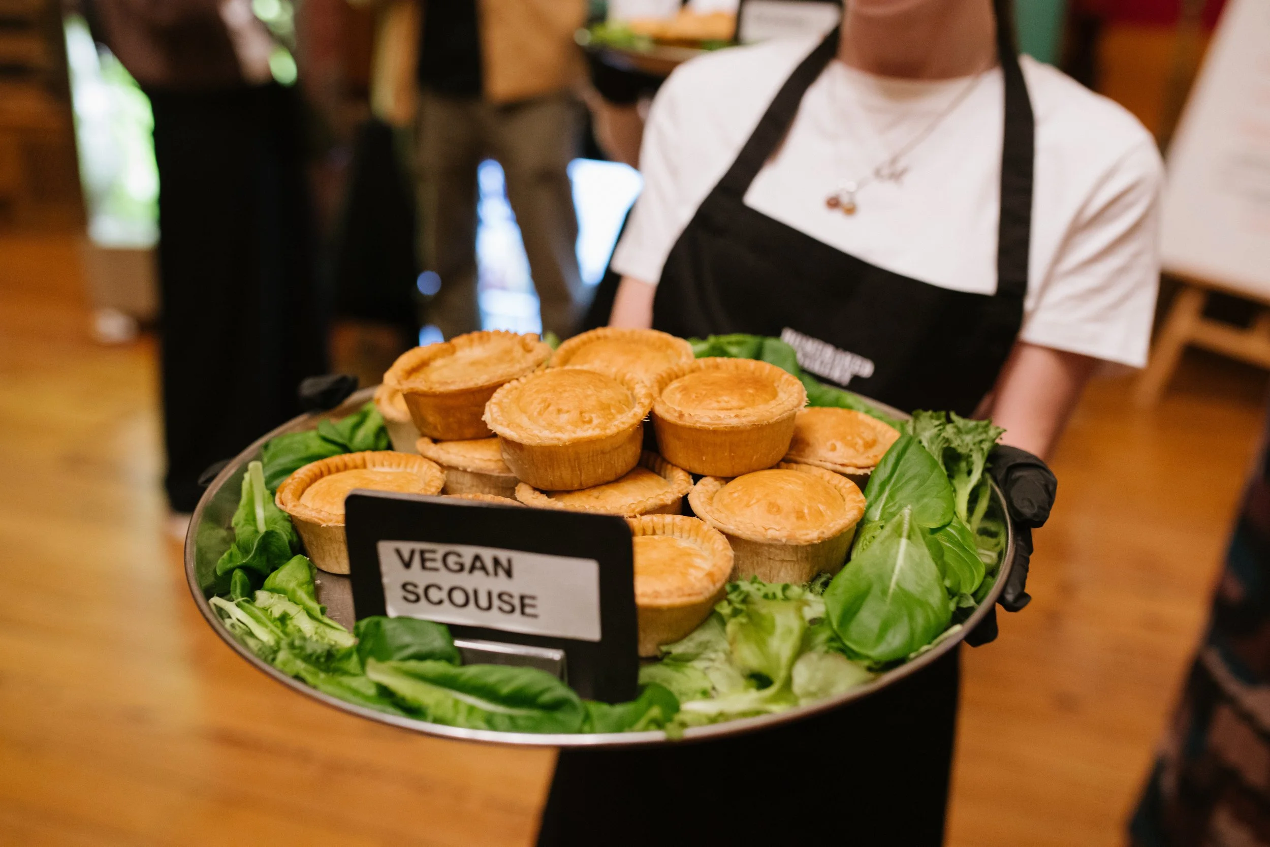 A person holds a large tray of vegan savory pies garnished with leafy greens at a restaurant or cafe.