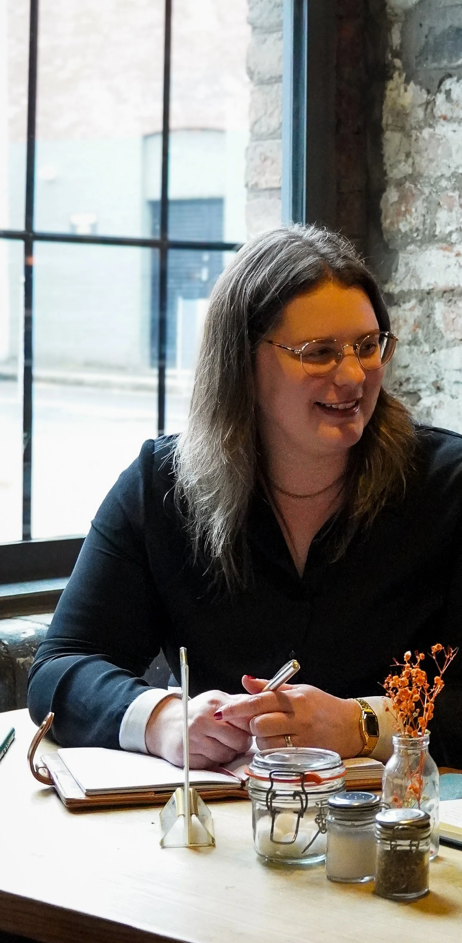 Rebecca sitting at a cafe table holding a pen in conversation and working from a notebook, behind are large windows.