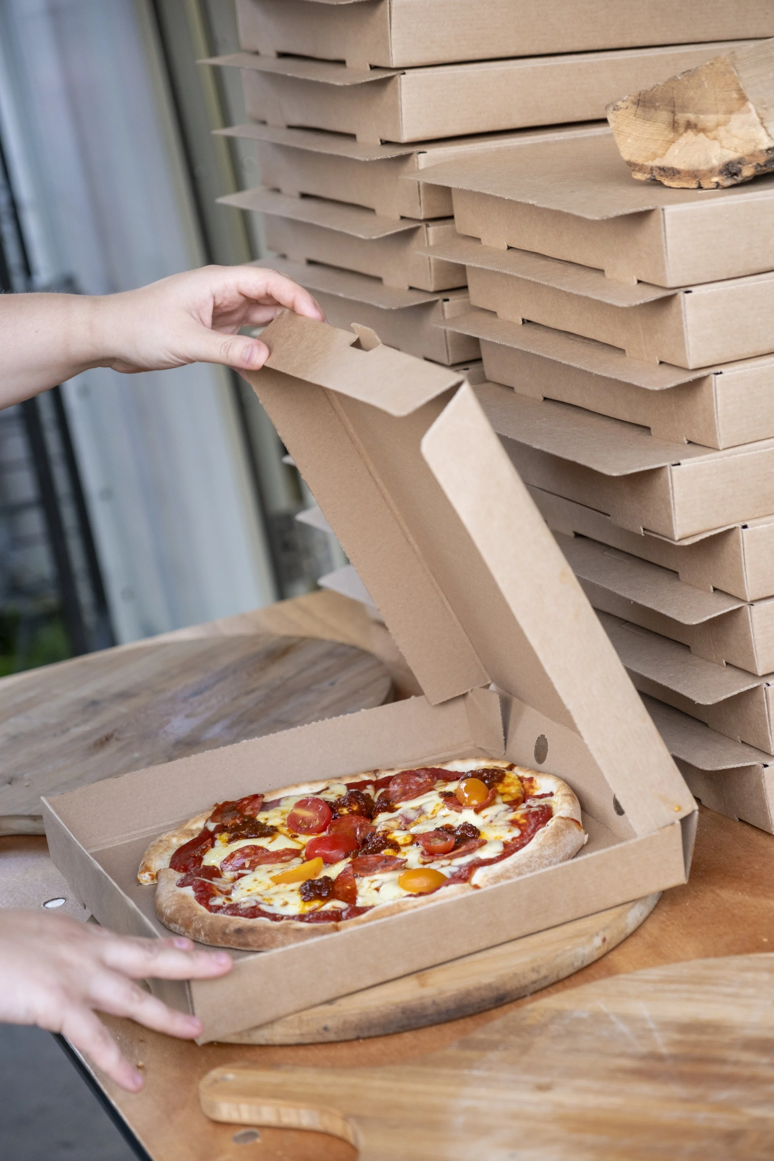 A person opening a pizza box with a cooked pizza topped with cherry tomatoes, cheese, and pepperoni on a wooden board, with stacked pizza boxes in the background.