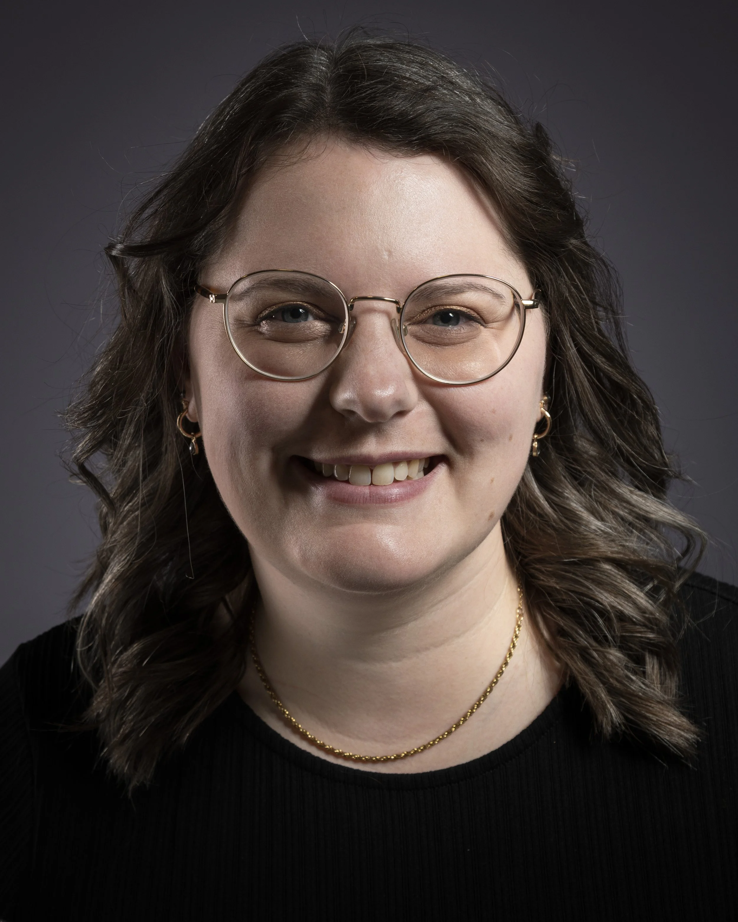 Photo of Rebecca, with shoulder-length brown hair, glasses, and earrings smiling against a dark gray background.