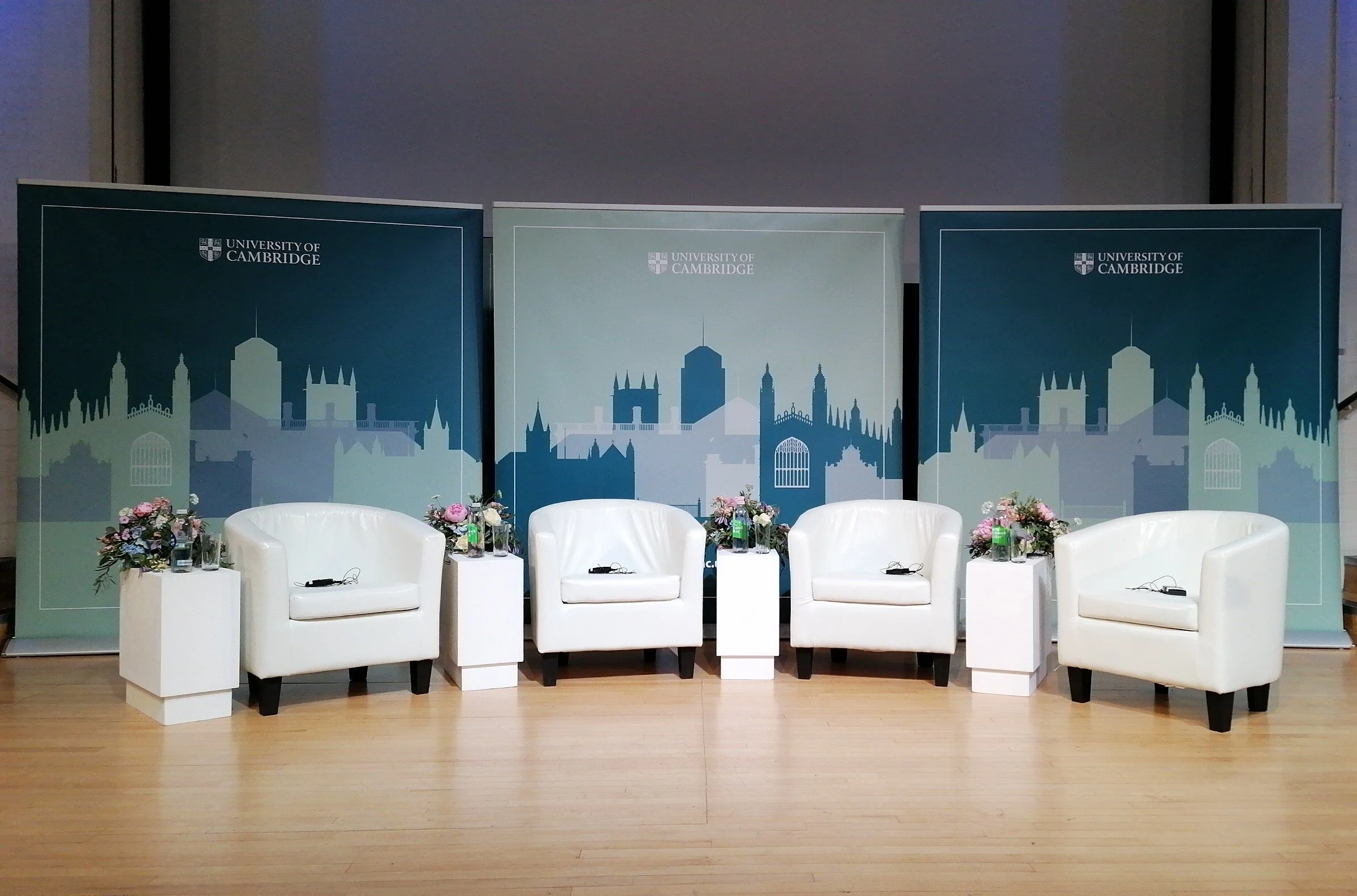 Conference setup with four white armchairs, small tables with flower arrangements, and University of Cambridge banners in the background.