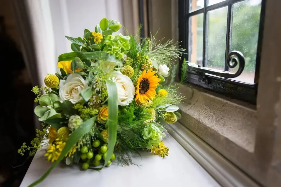 A bouquet of white roses, sunflowers, yellow and green flowers, and assorted greenery on a windowsill.